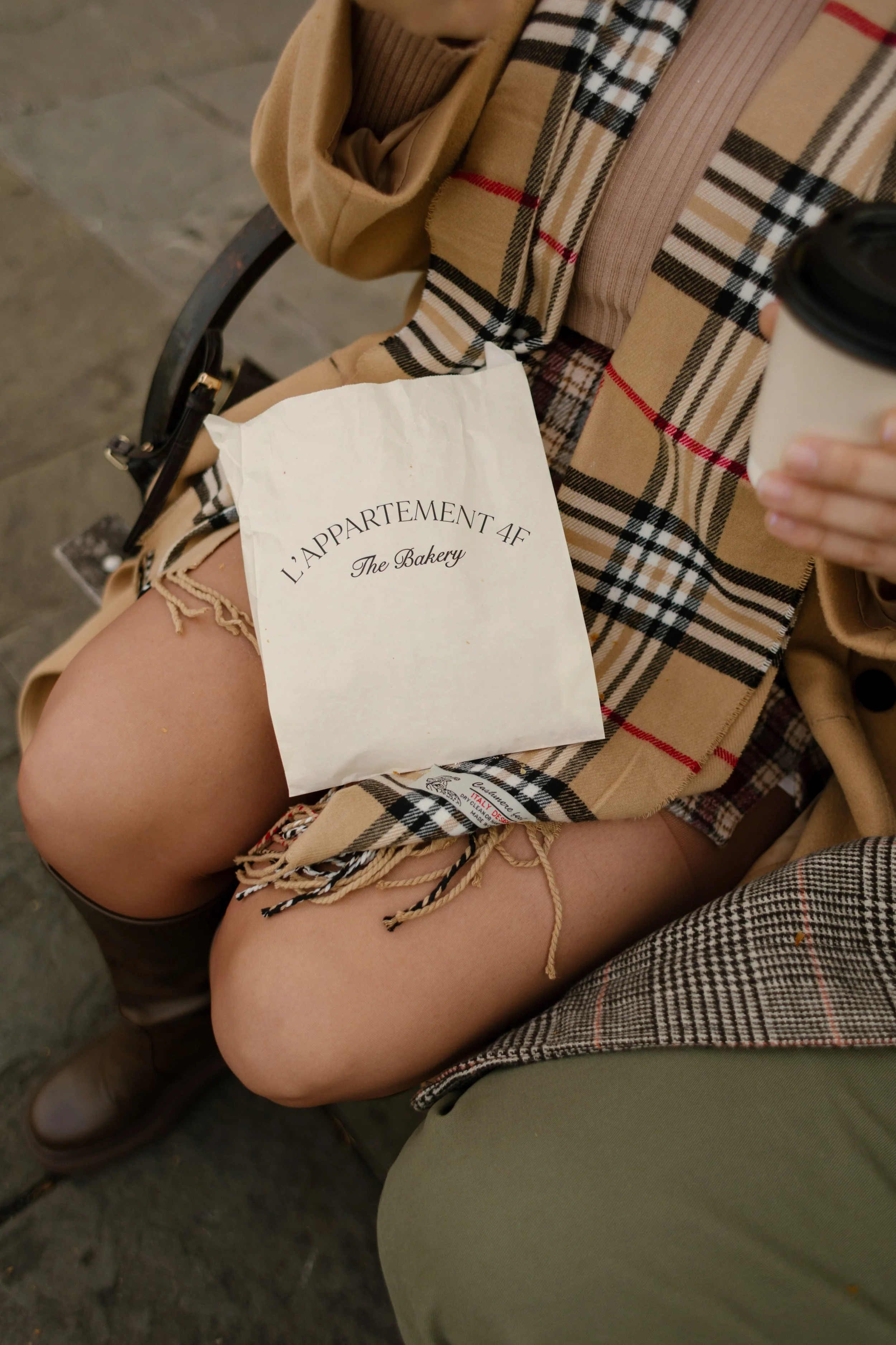 Close-up fall engagement detail with a bakery bag, coffee, plaid scarf, camel coat, and brown boots.