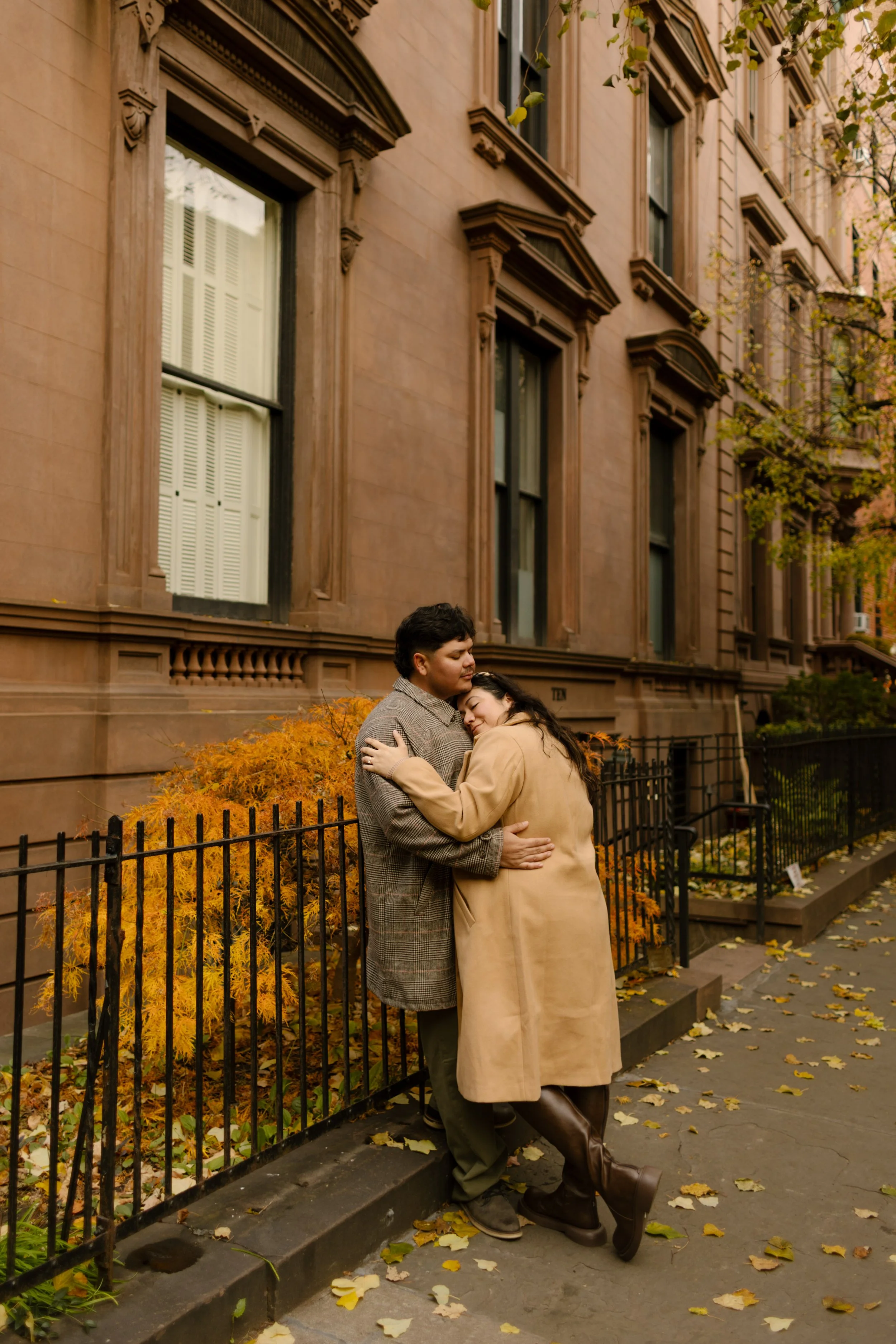 Brooklyn engagement photos of a couple embracing beside brownstones and golden fall foliage on a quiet neighborhood sidewalk.