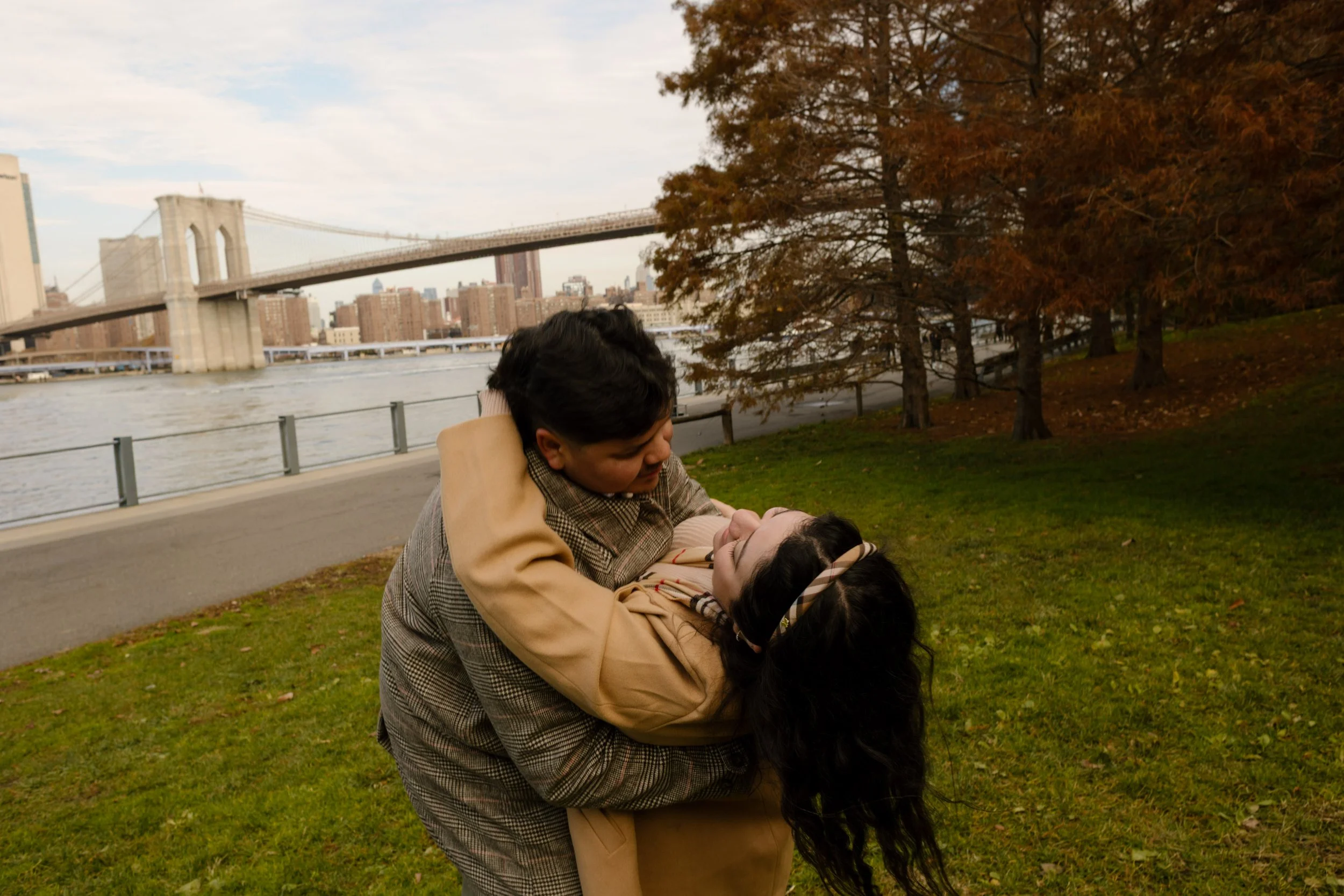 Playful fall engagement photo of a couple laughing and embracing near the Brooklyn Bridge waterfront.