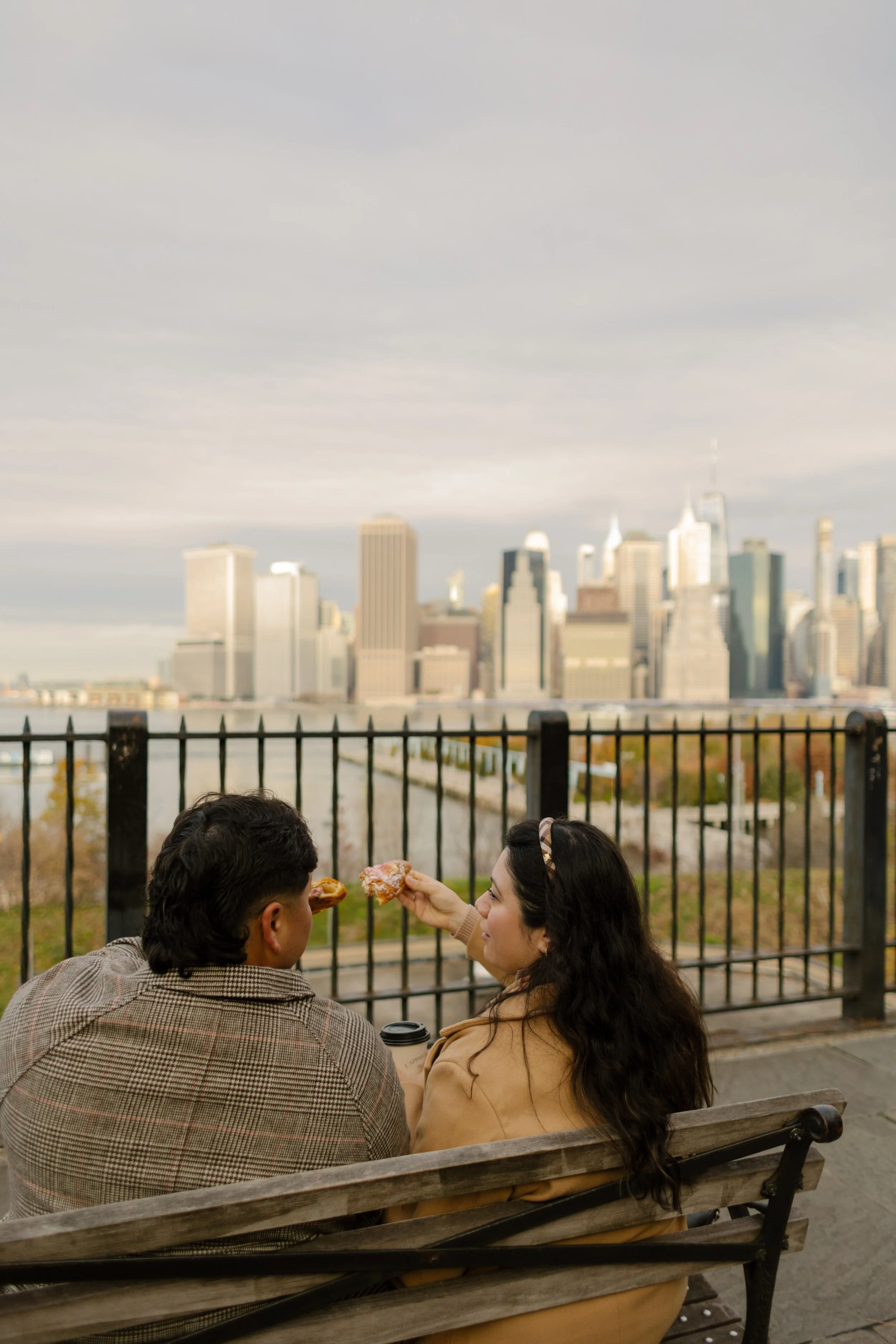 1. Couple sharing pastries and coffee on a bench overlooking the Manhattan skyline during a cozy fall engagement session.