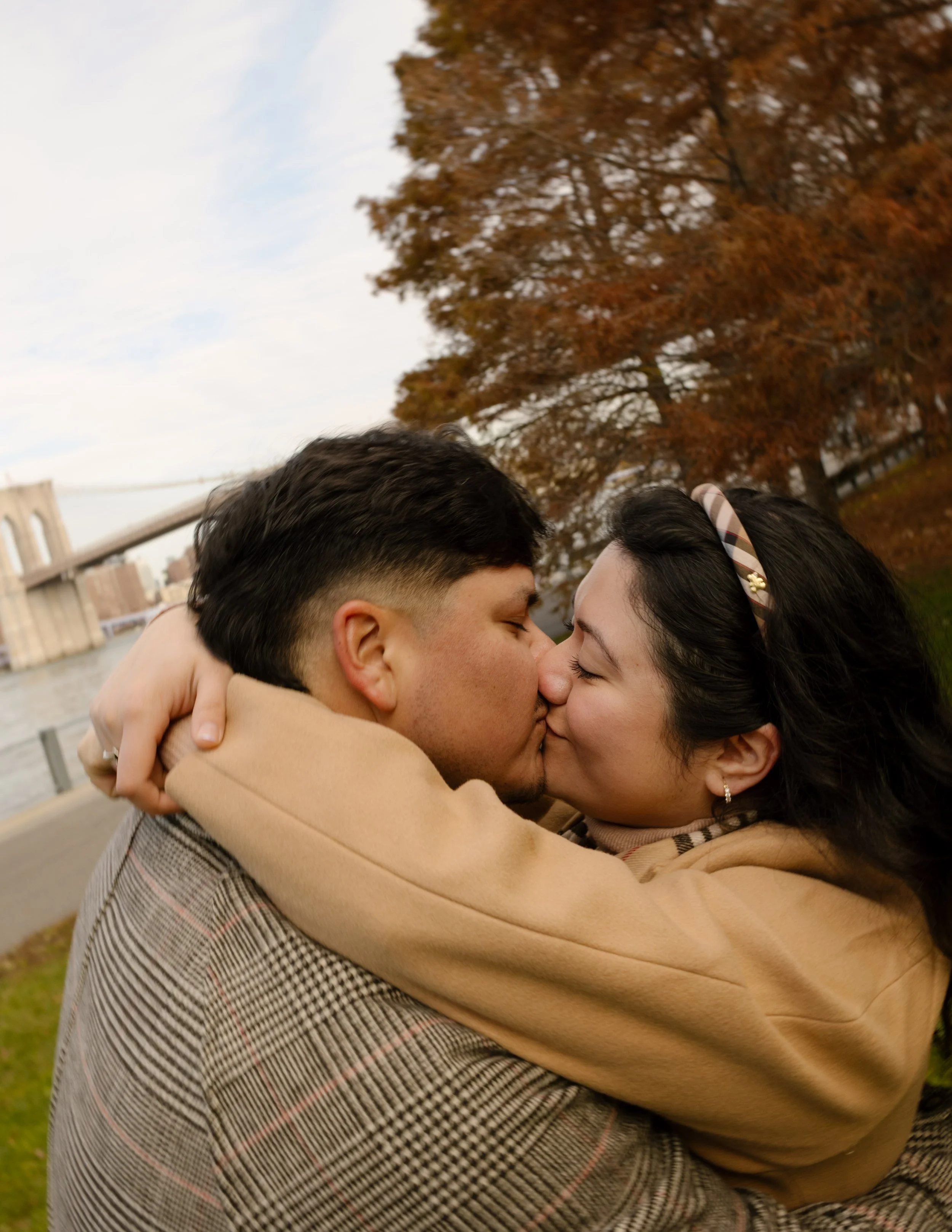 Close-up of an engaged couple kissing near the Brooklyn waterfront with colorful fall trees in the background.