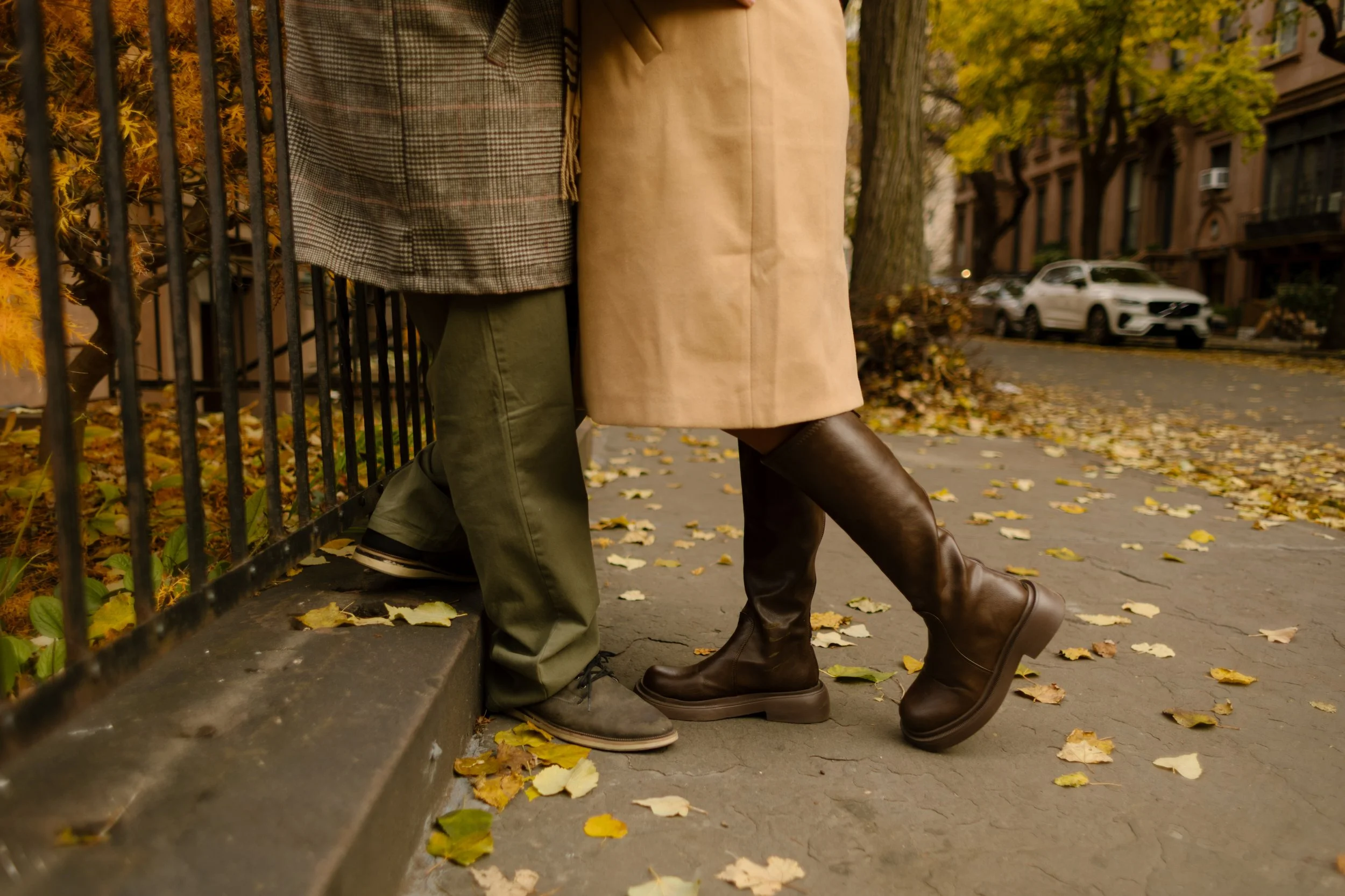 Close-up of a couple’s fall boots together on a leaf-covered sidewalk lined with Brooklyn brownstones.