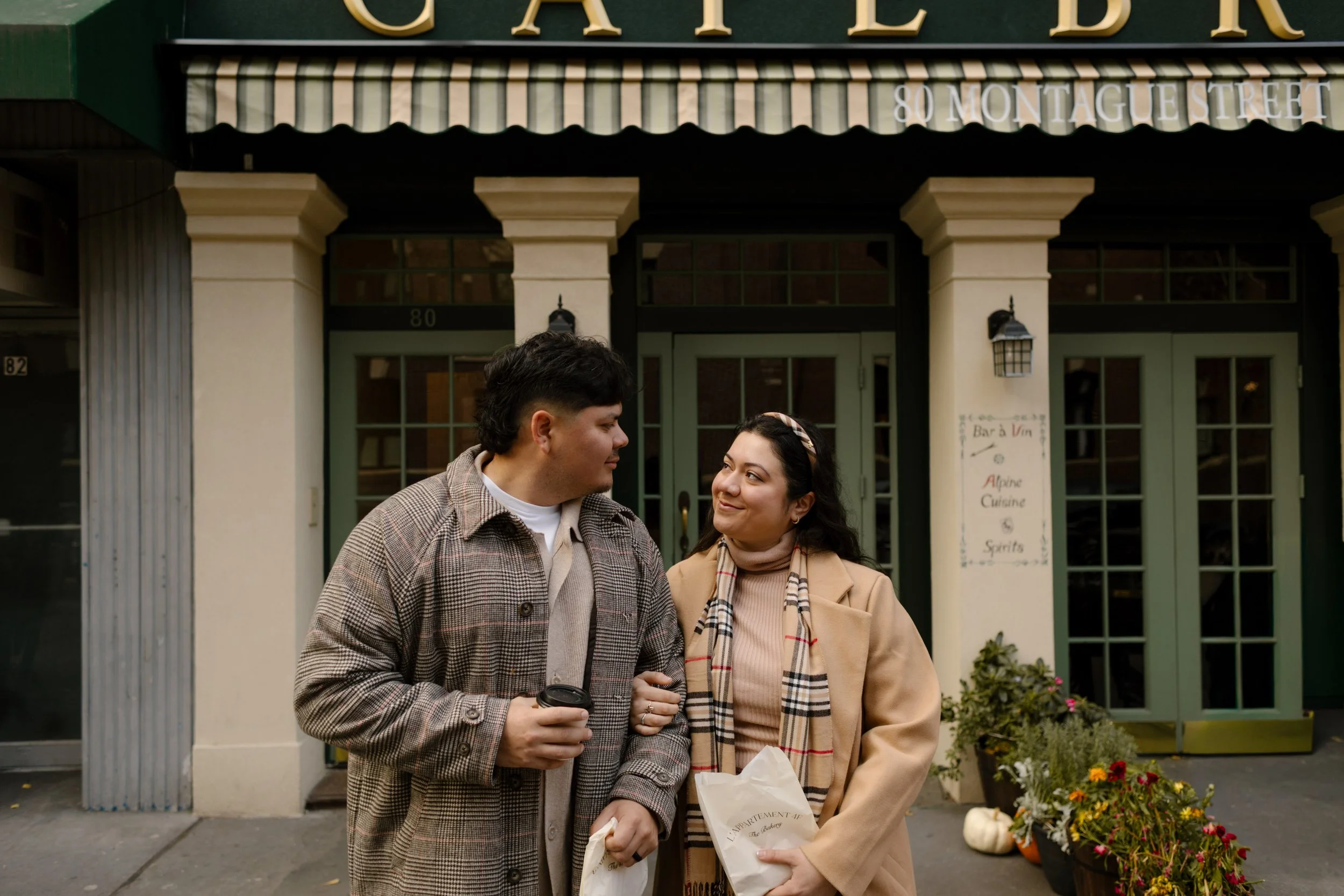 Brooklyn engagement photos of a couple walking together with coffee and pastries outside a neighborhood café.