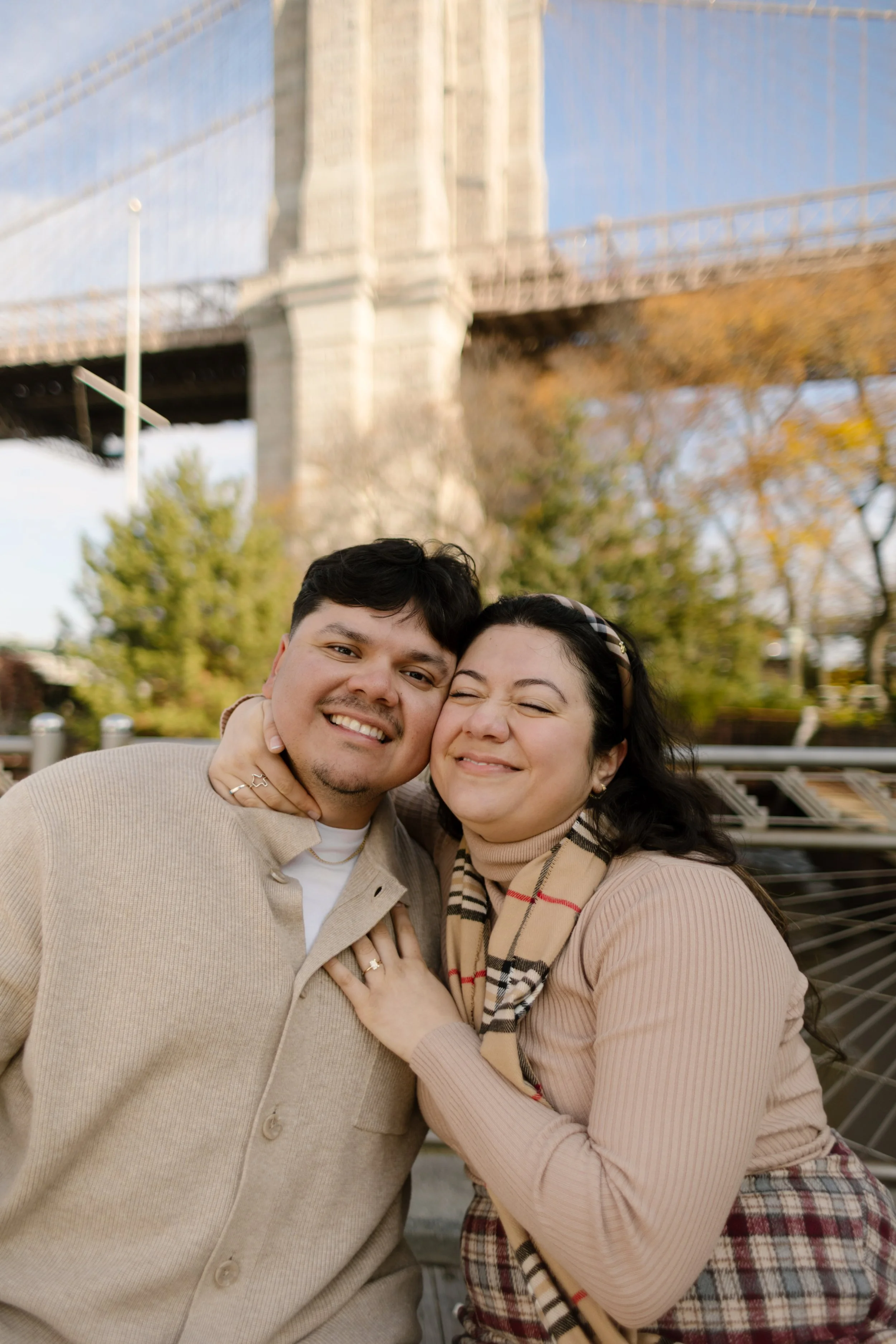 1. Close-up fall engagement portrait of a smiling couple cuddling together with the Brooklyn Bridge in the background.