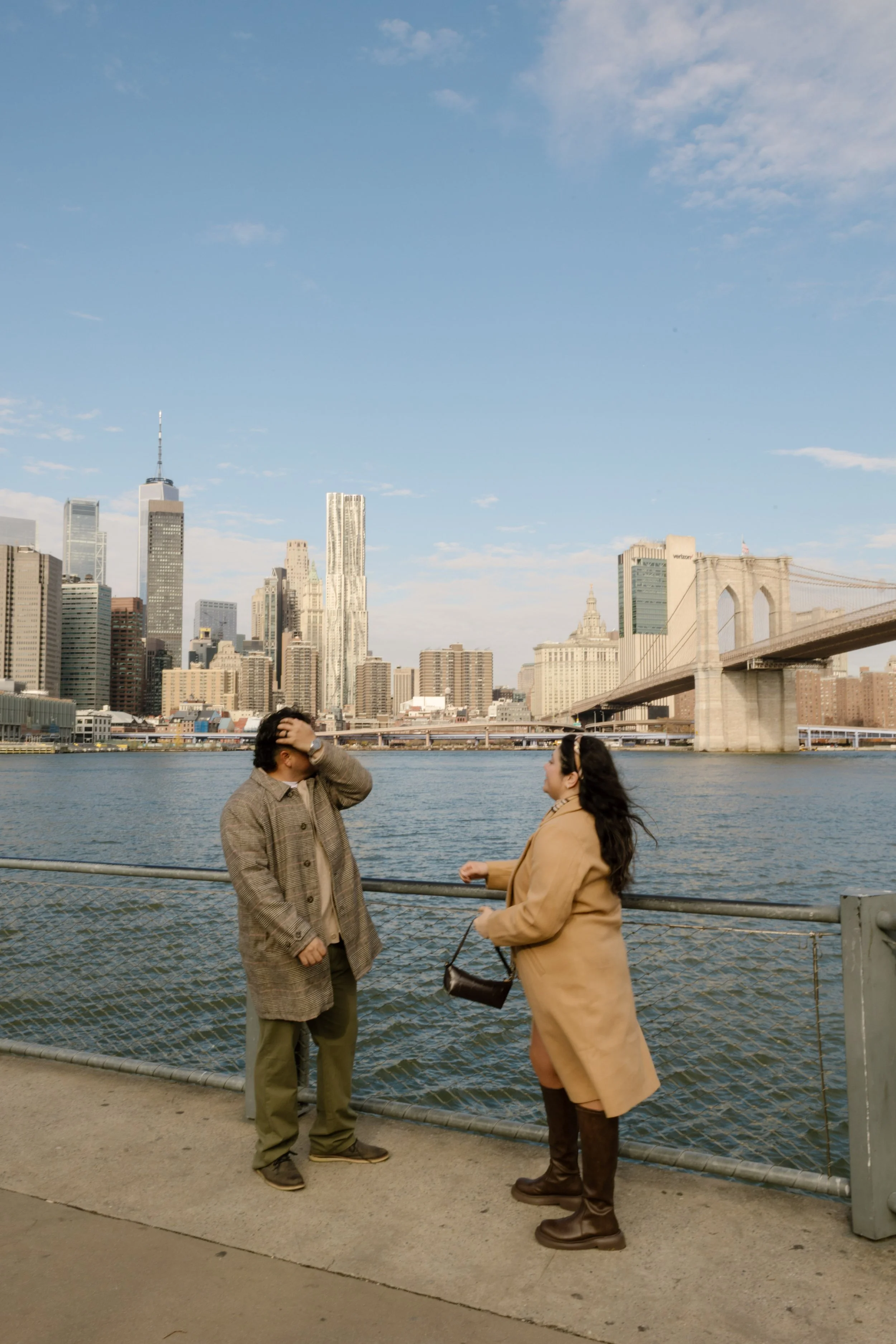 Brooklyn engagement photos of a couple laughing along the waterfront with the Brooklyn Bridge and Manhattan skyline behind them.