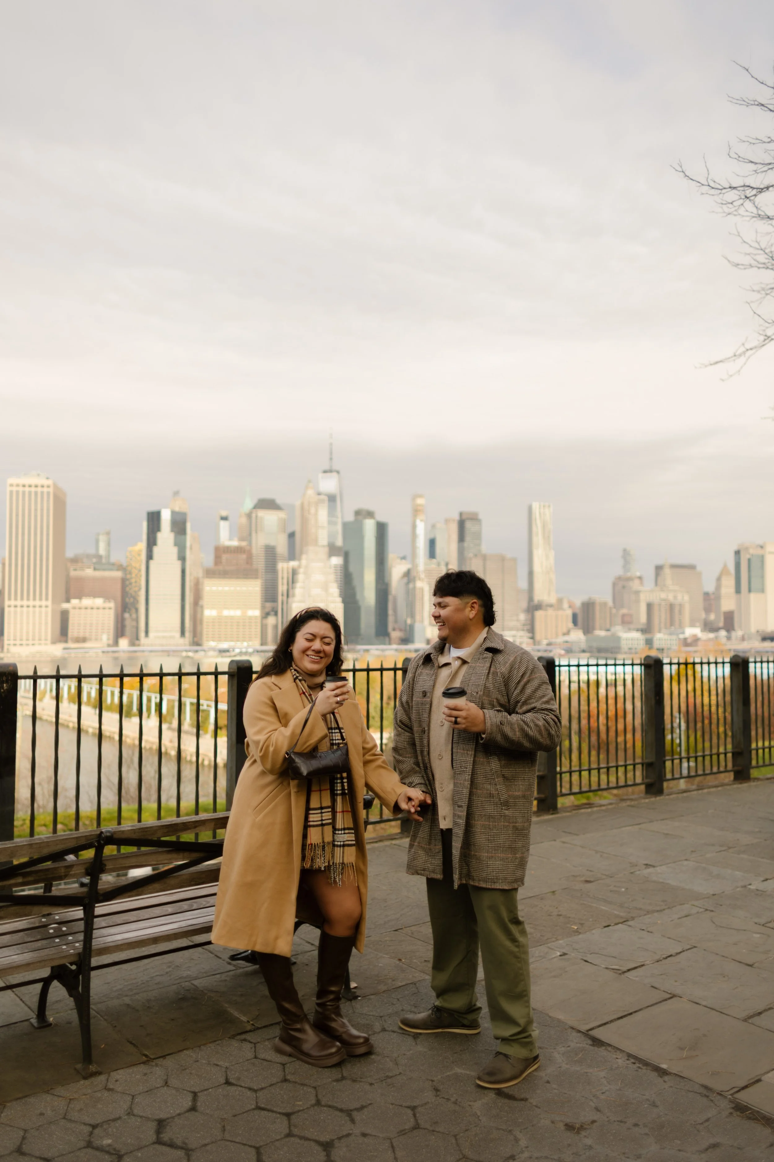 Brooklyn engagement photos of a couple laughing with coffee while overlooking the Manhattan skyline on a fall day.