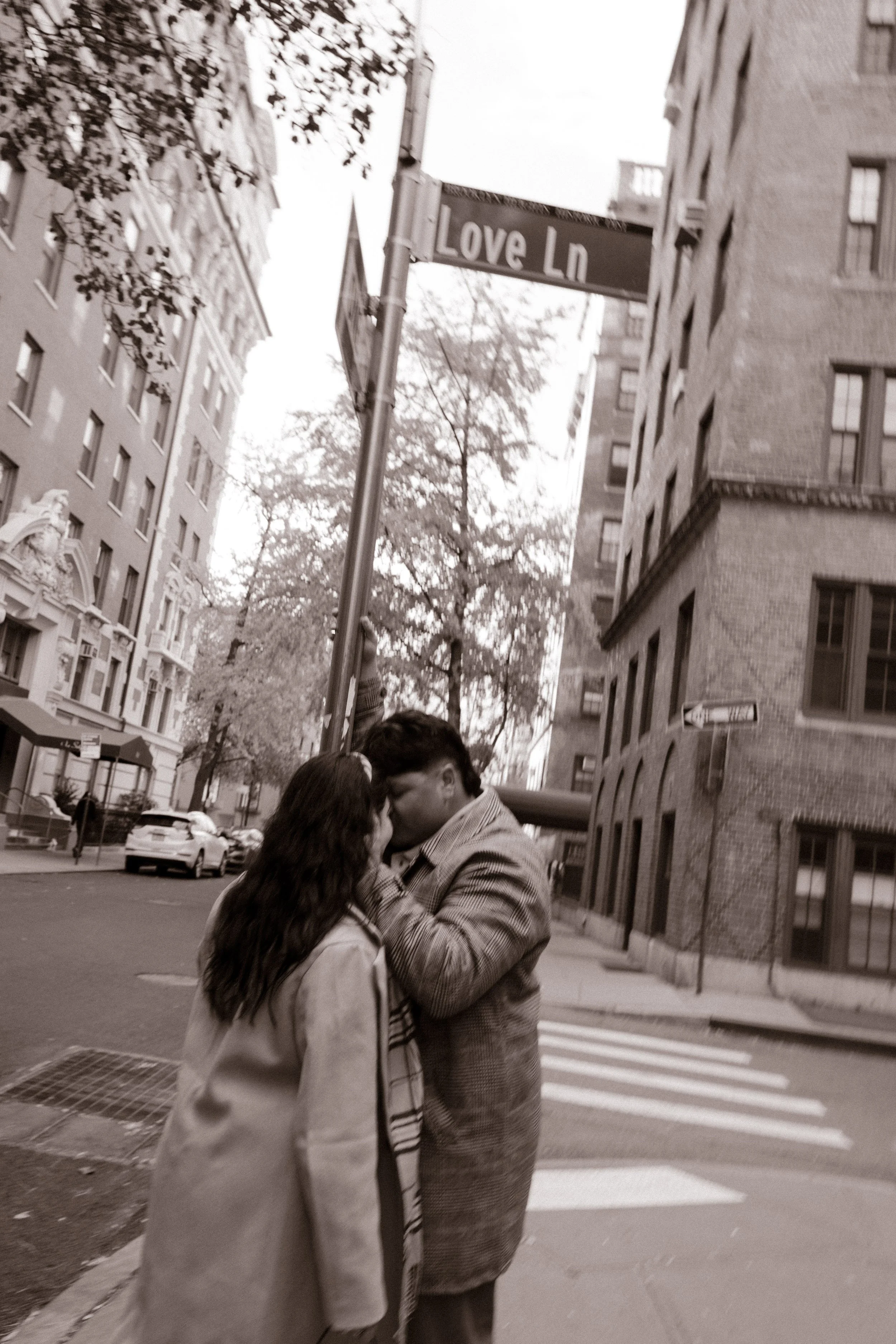 Black-and-white candid photo of a couple kissing on a Brooklyn street beneath a Love Ln street sign.