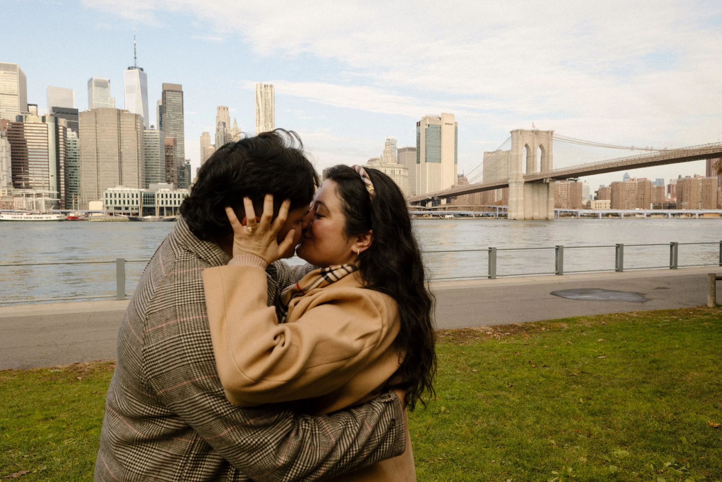 Brooklyn engagement photos of a couple kissing by the East River with the Brooklyn Bridge and Manhattan skyline in the background.