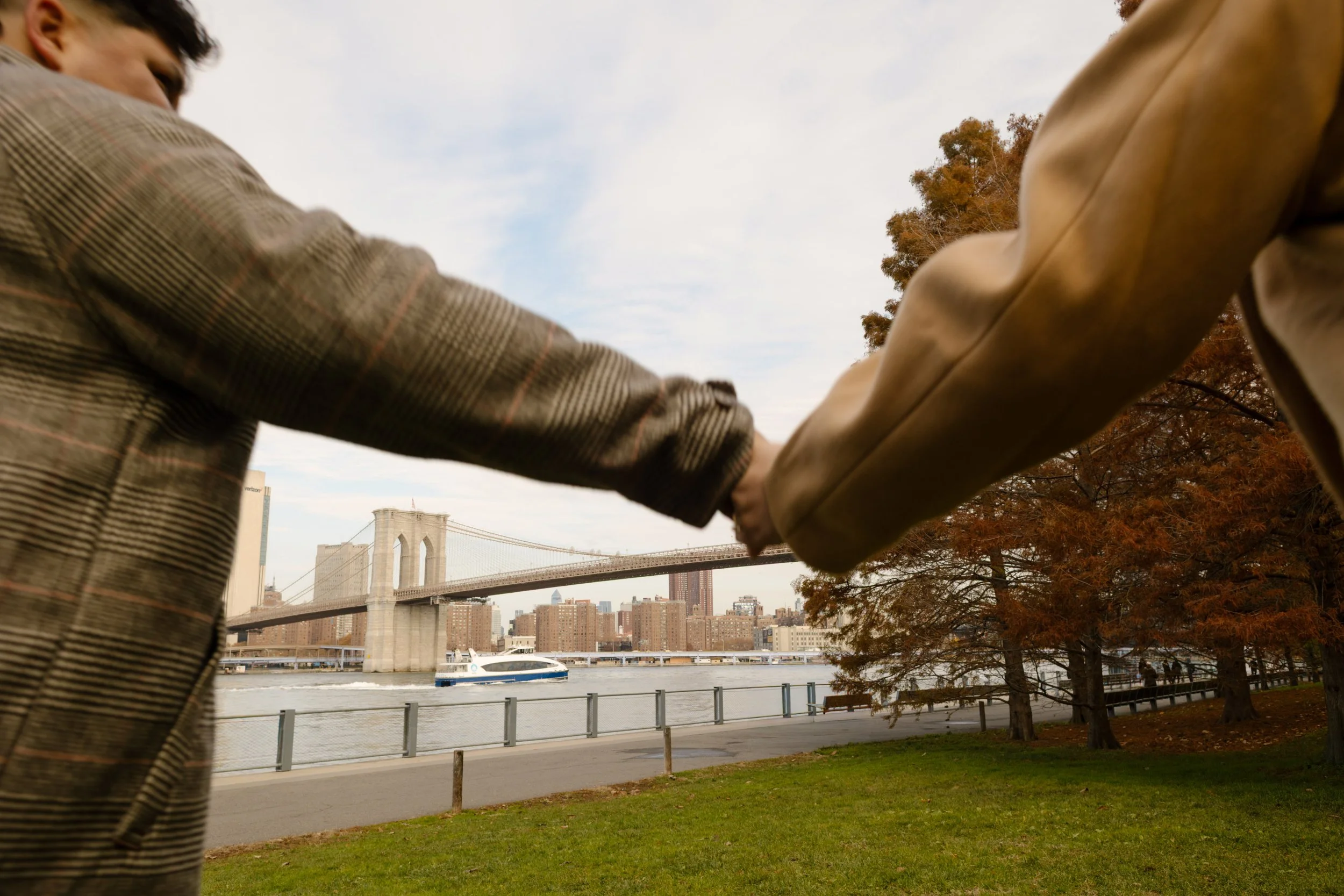 Brooklyn engagement photos of a couple holding hands by the waterfront with the Brooklyn Bridge and fall trees in the background.