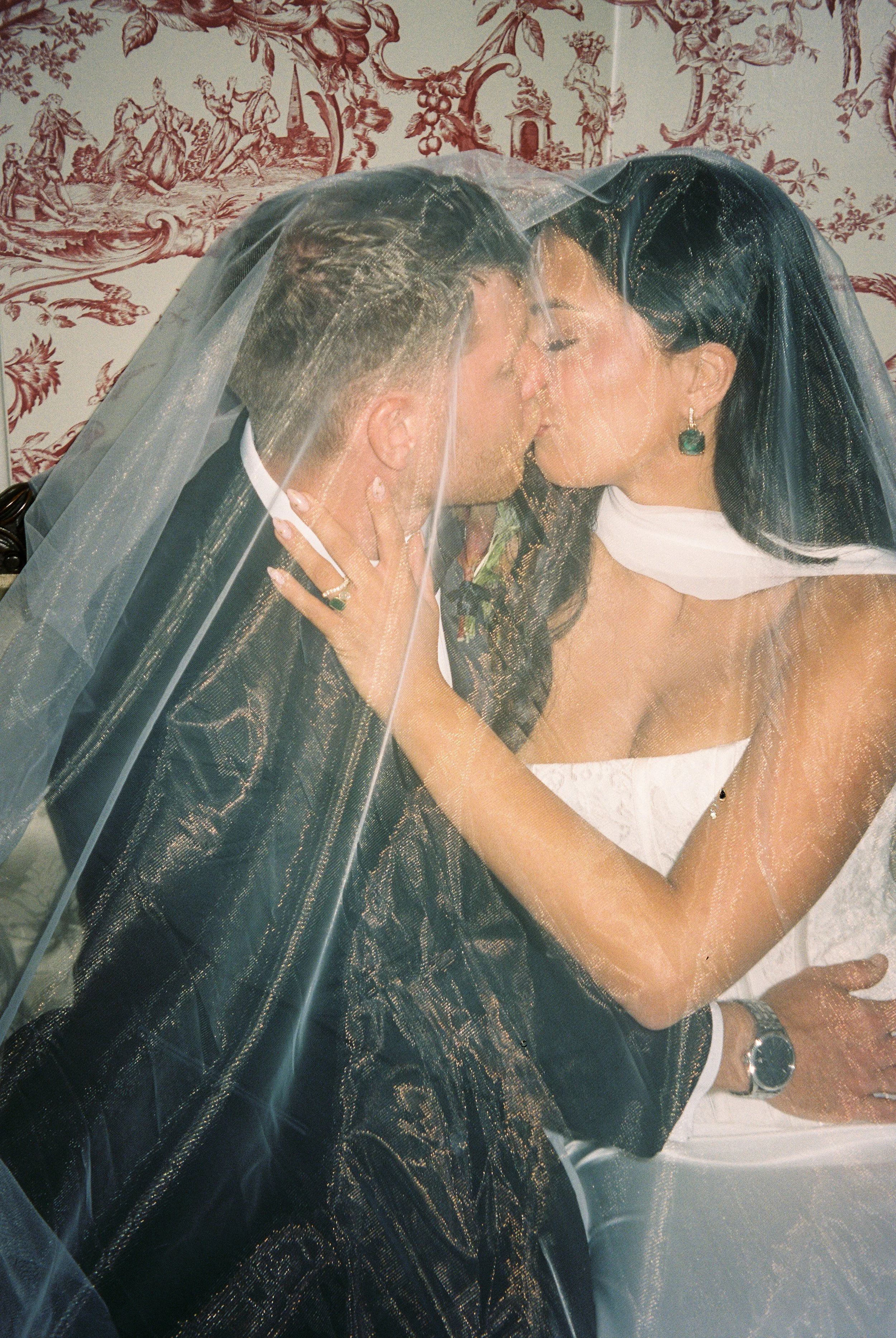 Romantic film wedding photo of a bride and groom kissing beneath her veil against vintage patterned wallpaper.