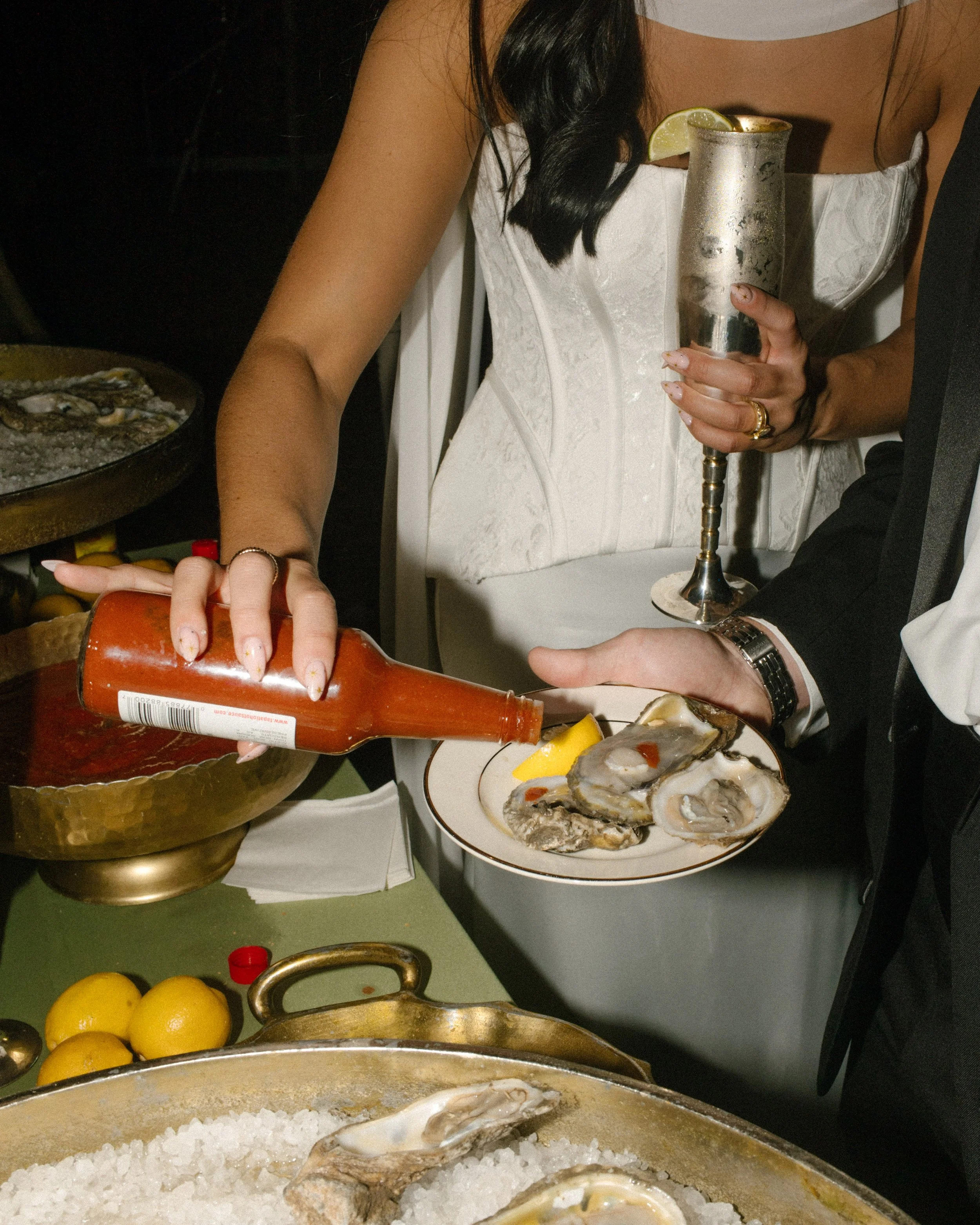 Bride in a white corset wedding dress pouring hot sauce onto oysters during her wedding reception.