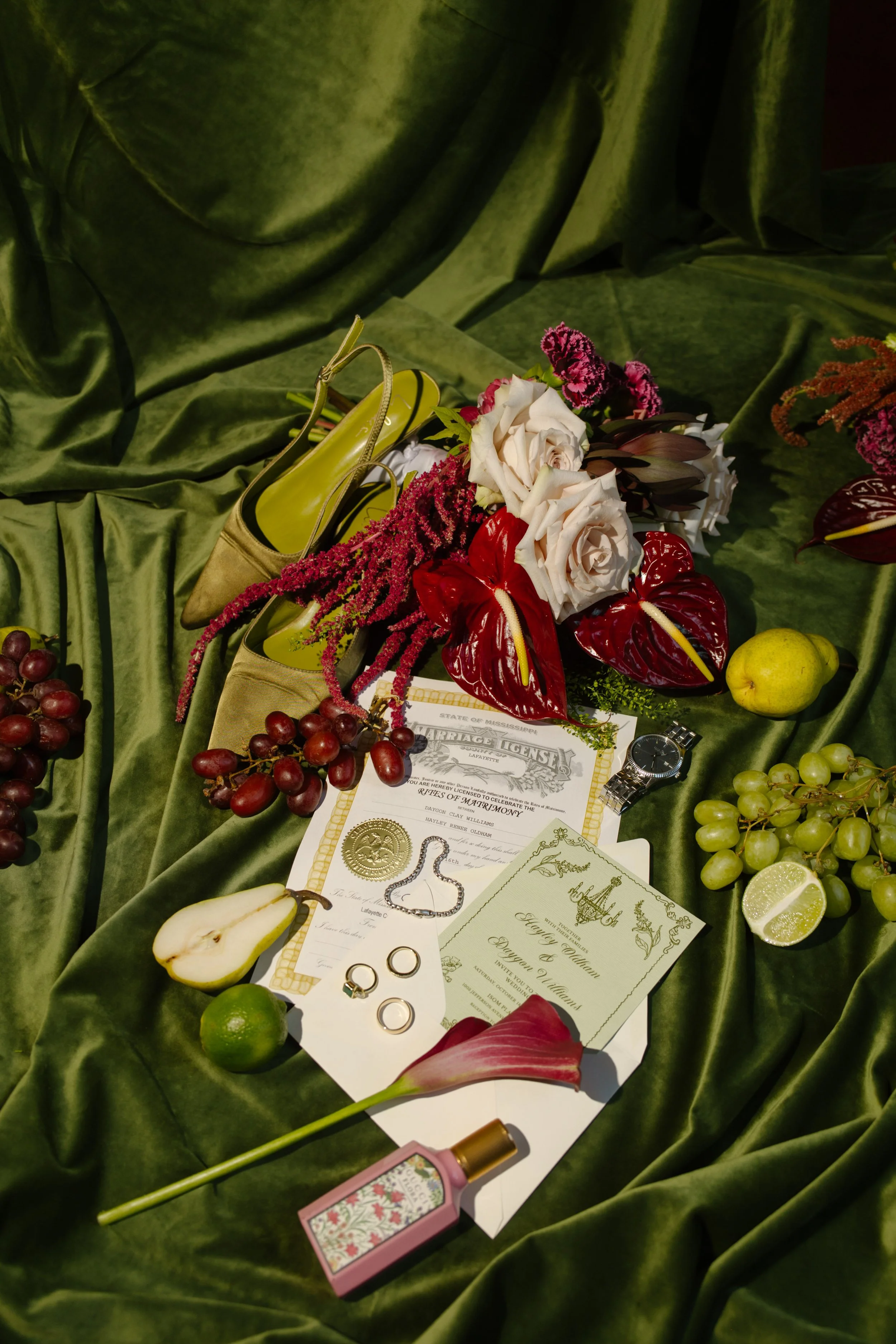 Wedding flat lay featuring moody wedding colors with olive green velvet, burgundy florals, ivory roses, fruit, and bridal details.