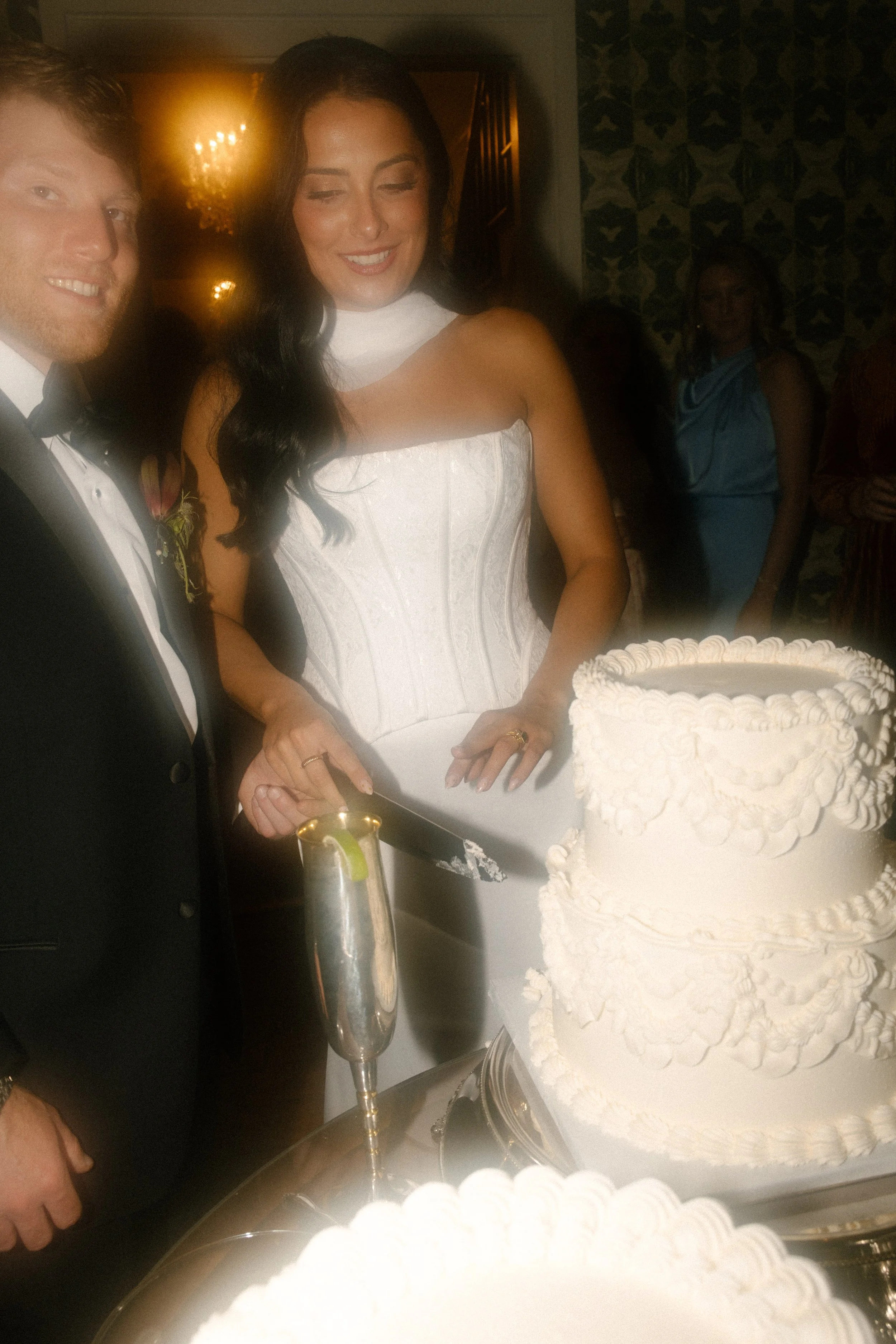 Bride and groom cutting a vintage white wedding cake in a dimly lit reception with moody wedding colors and warm lighting.