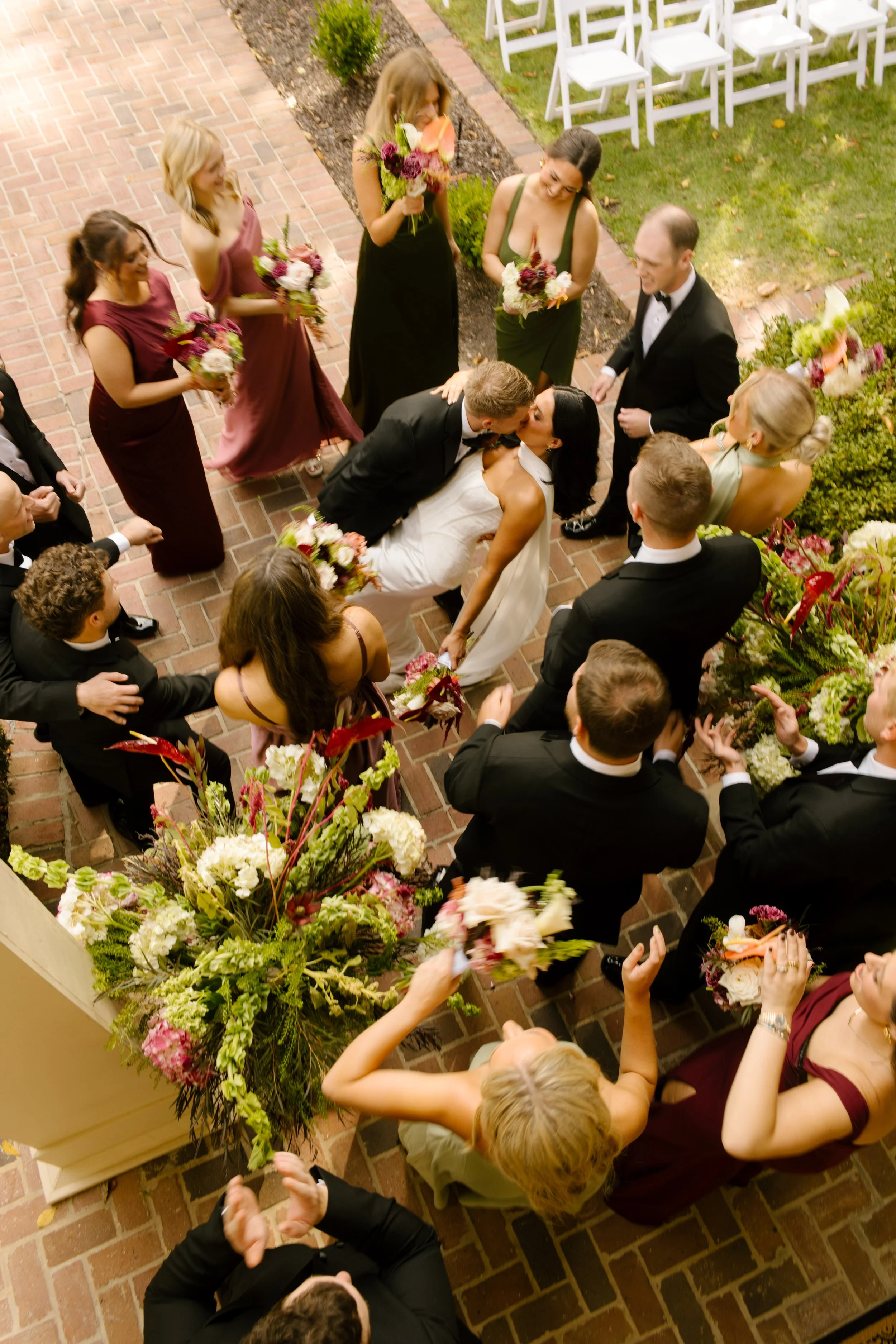 Overhead wedding party photo of the bride and groom kissing surrounded by bridesmaids, groomsmen, and colorful bouquets.