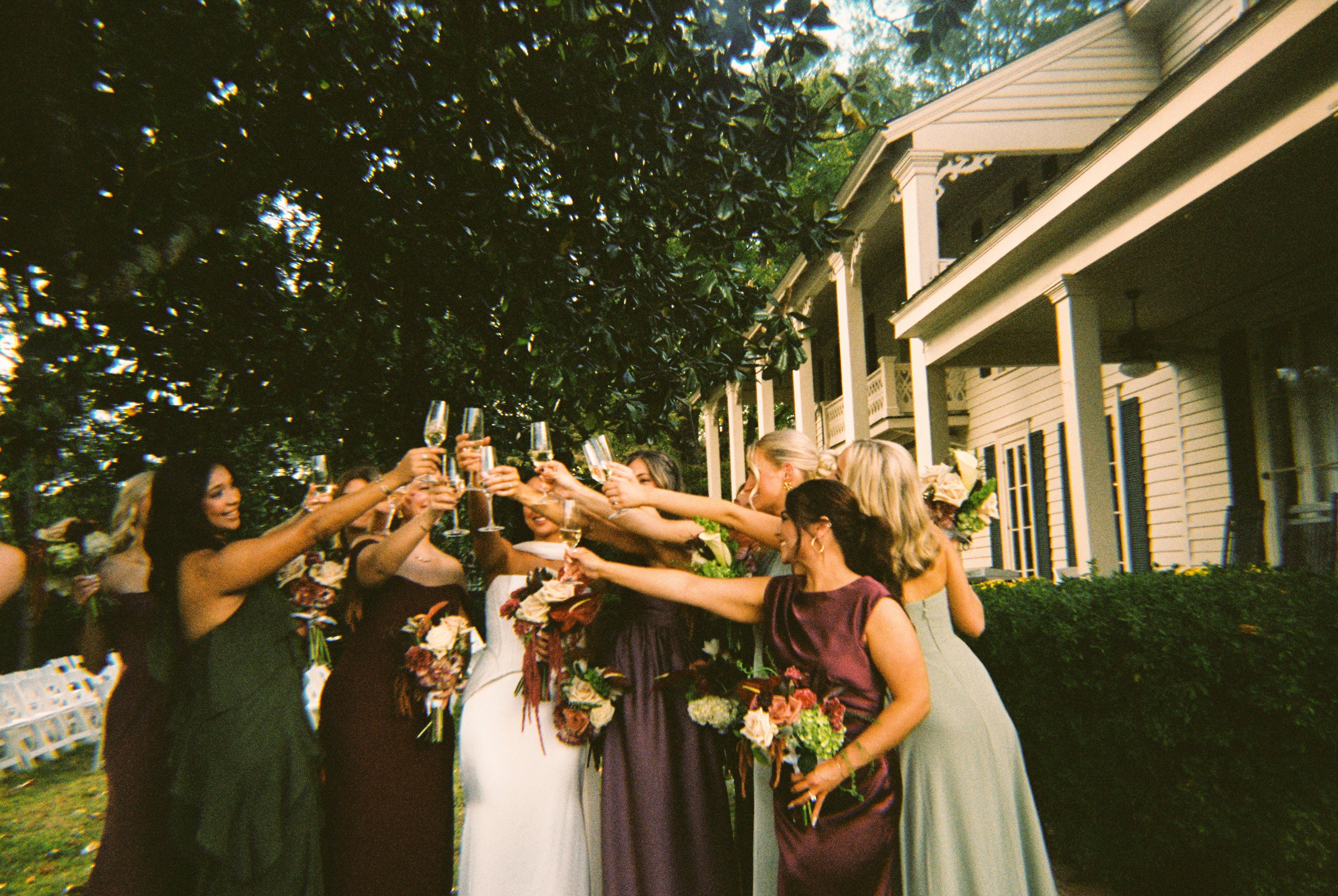 Bridesmaids raising champagne glasses in a celebratory toast, wearing dresses in moody wedding colors of olive green, burgundy, and mauve.