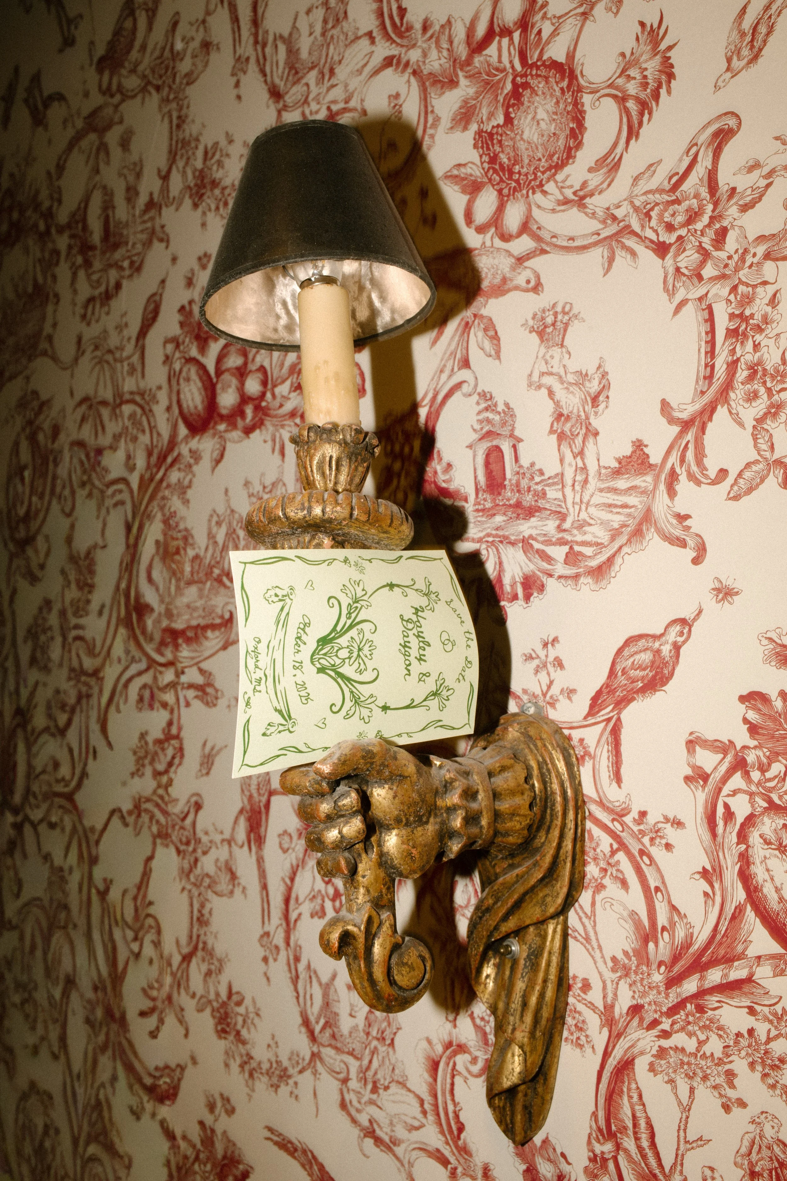 Vintage gold wall sconce holding wedding stationery against romantic red toile wallpaper.