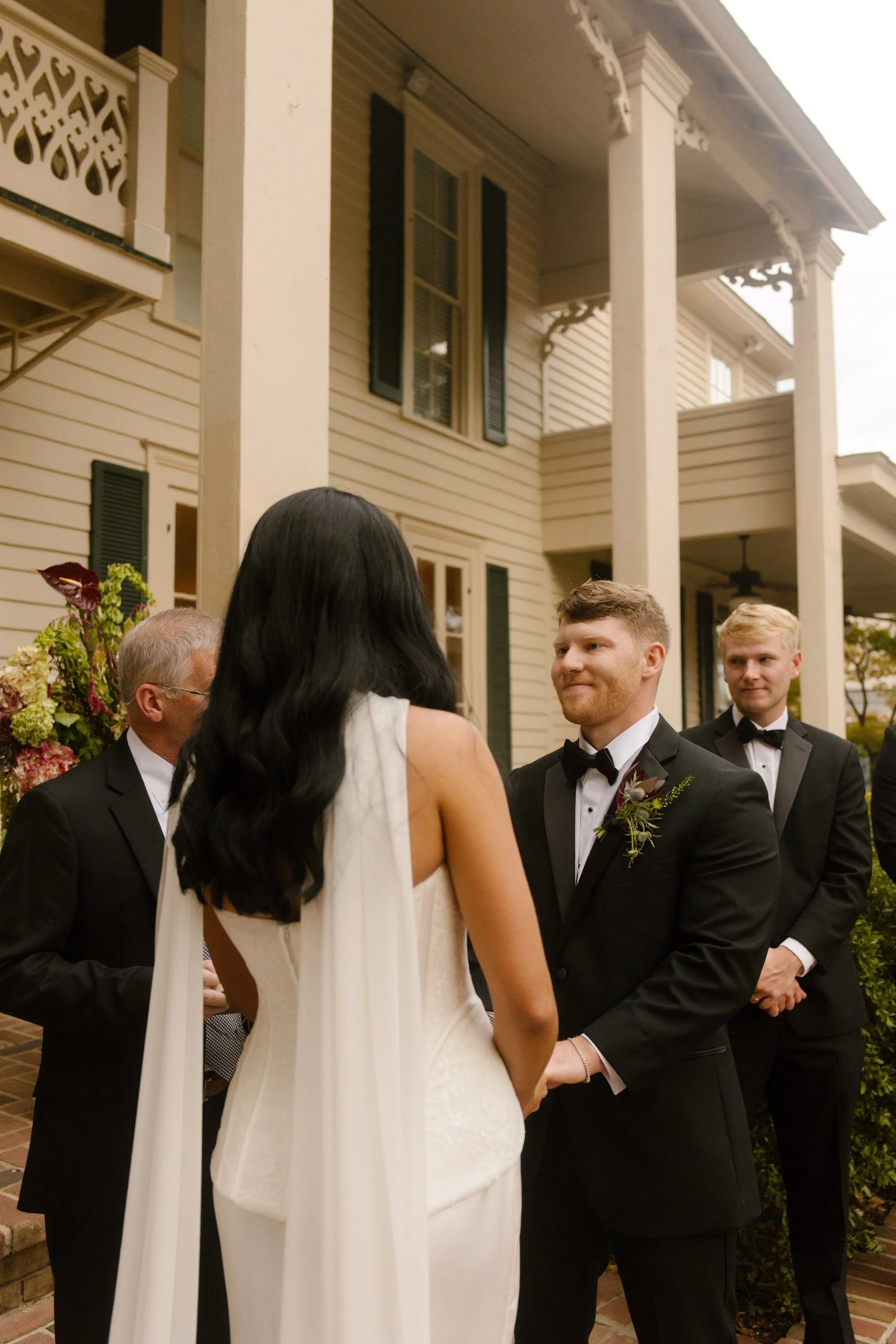 Groom smiling at his bride during their outdoor wedding ceremony in front of a historic white house.