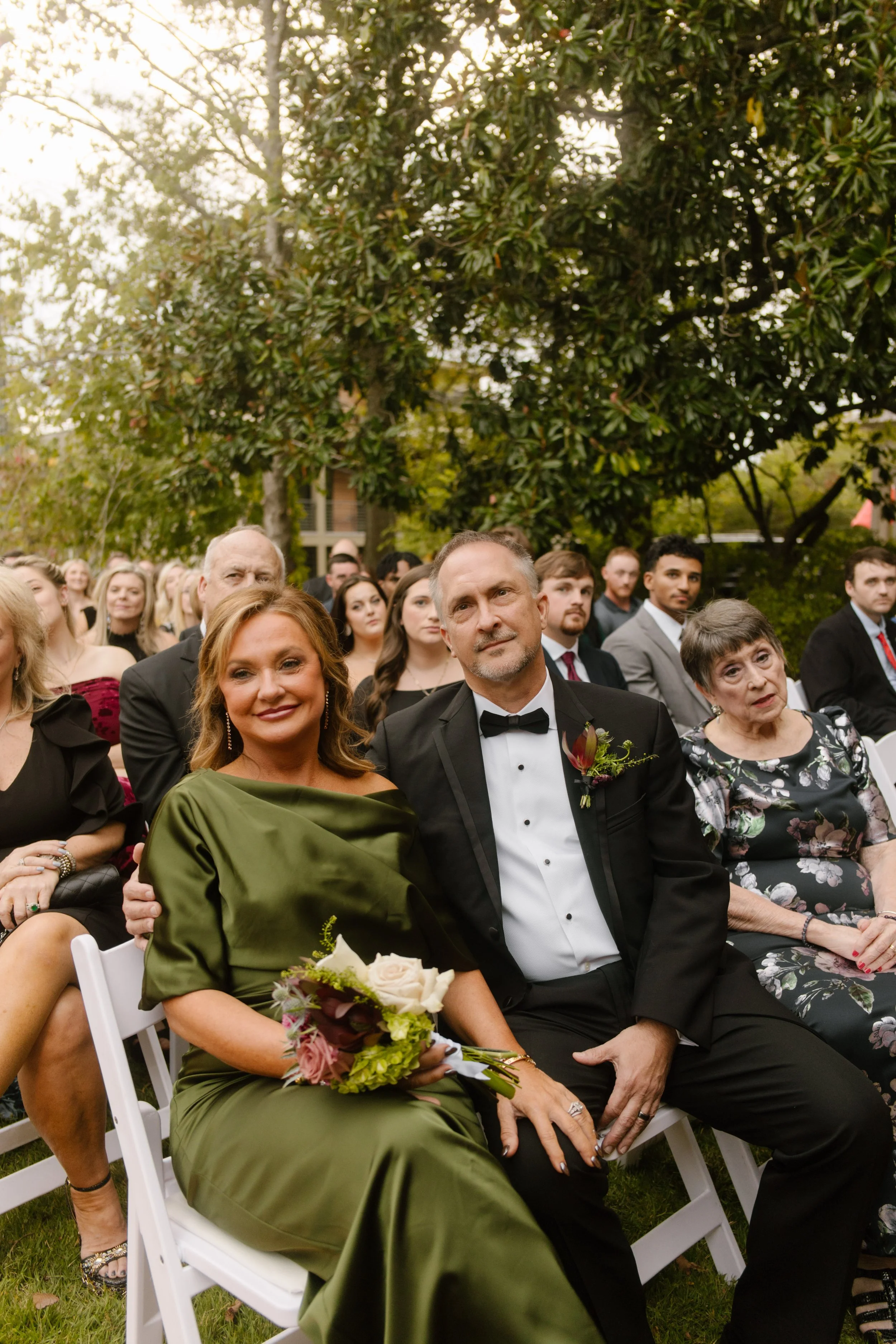 Wedding guests seated at an outdoor ceremony, featuring moody wedding colors with an olive green satin dress and burgundy floral accents.
