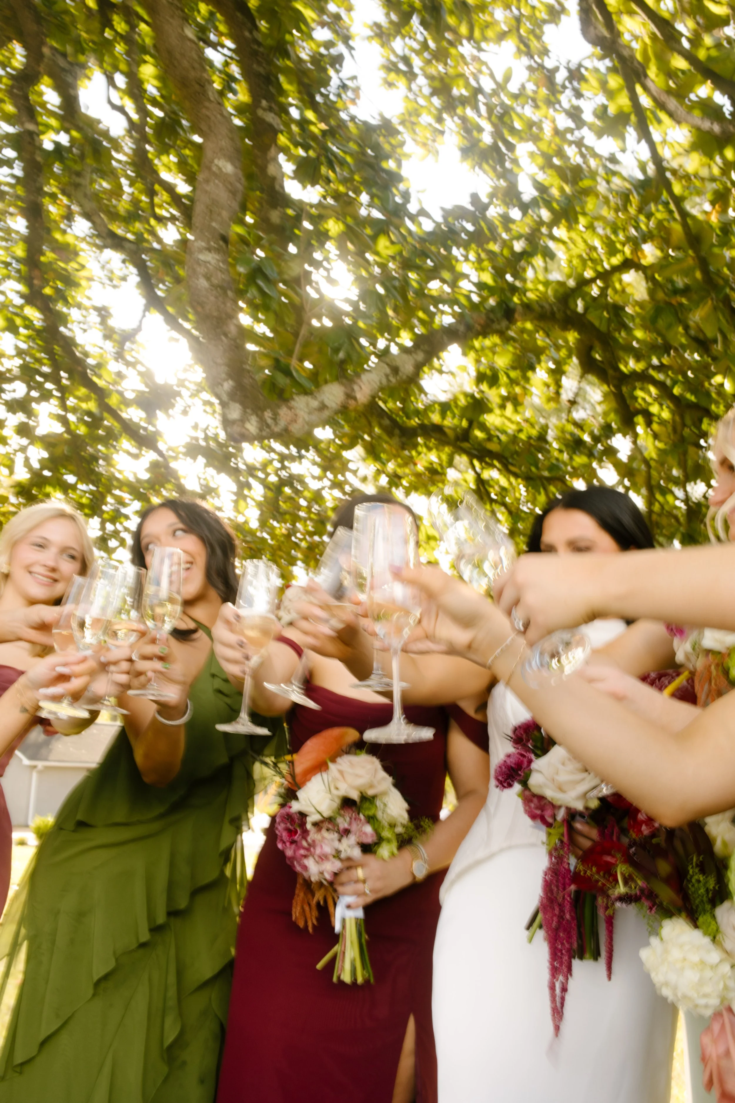 Bride and bridesmaids raising champagne glasses in a joyful toast, wearing moody wedding colors of olive green and burgundy with rich floral bouquets.