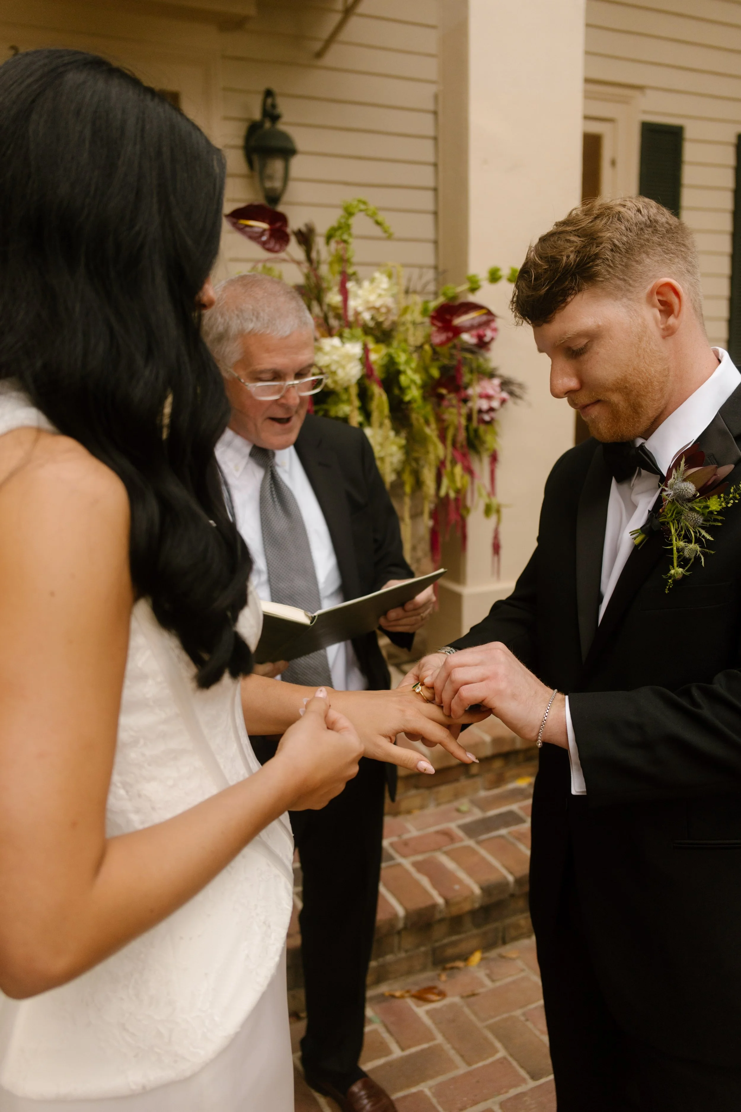 Groom placing a wedding ring on the bride's finger during an outdoor ceremony with moody wedding colors in the burgundy floral arrangements.