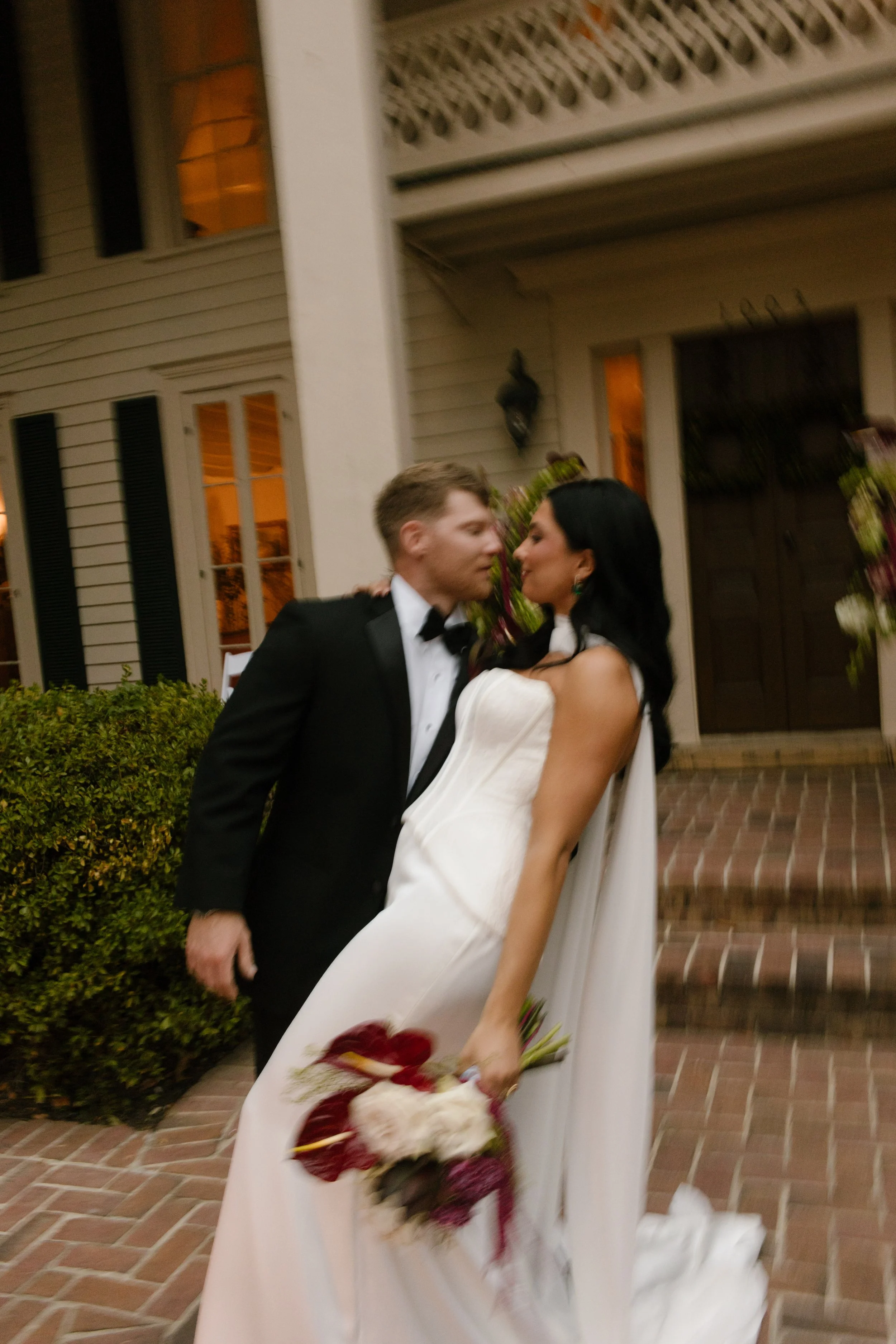 Candid bride and groom portraits featuring moody wedding colors in a deep burgundy and ivory bridal bouquet outside a historic house.