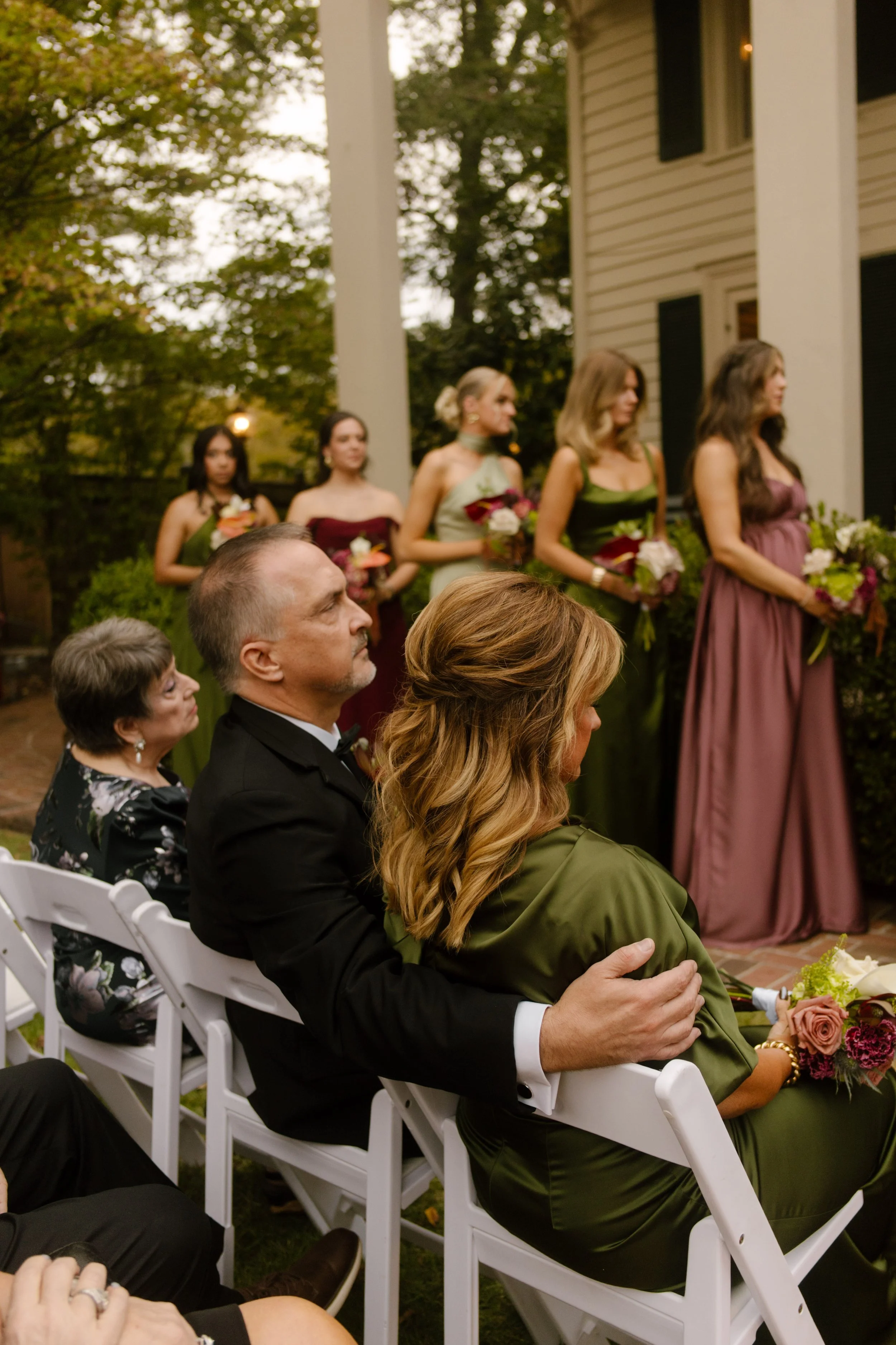 Wedding guests seated at an outdoor ceremony with bridesmaids wearing olive green, burgundy, and mauve dresses.