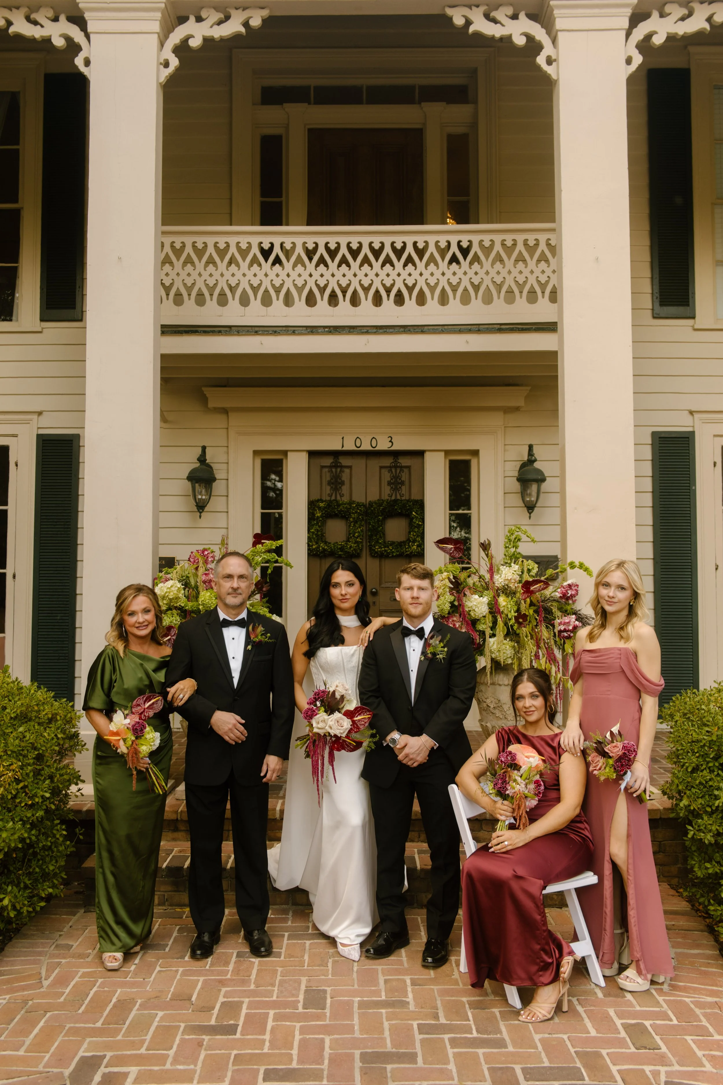 Wedding party portrait outside a historic venue featuring moody wedding colors in olive green and burgundy attire with dramatic florals.