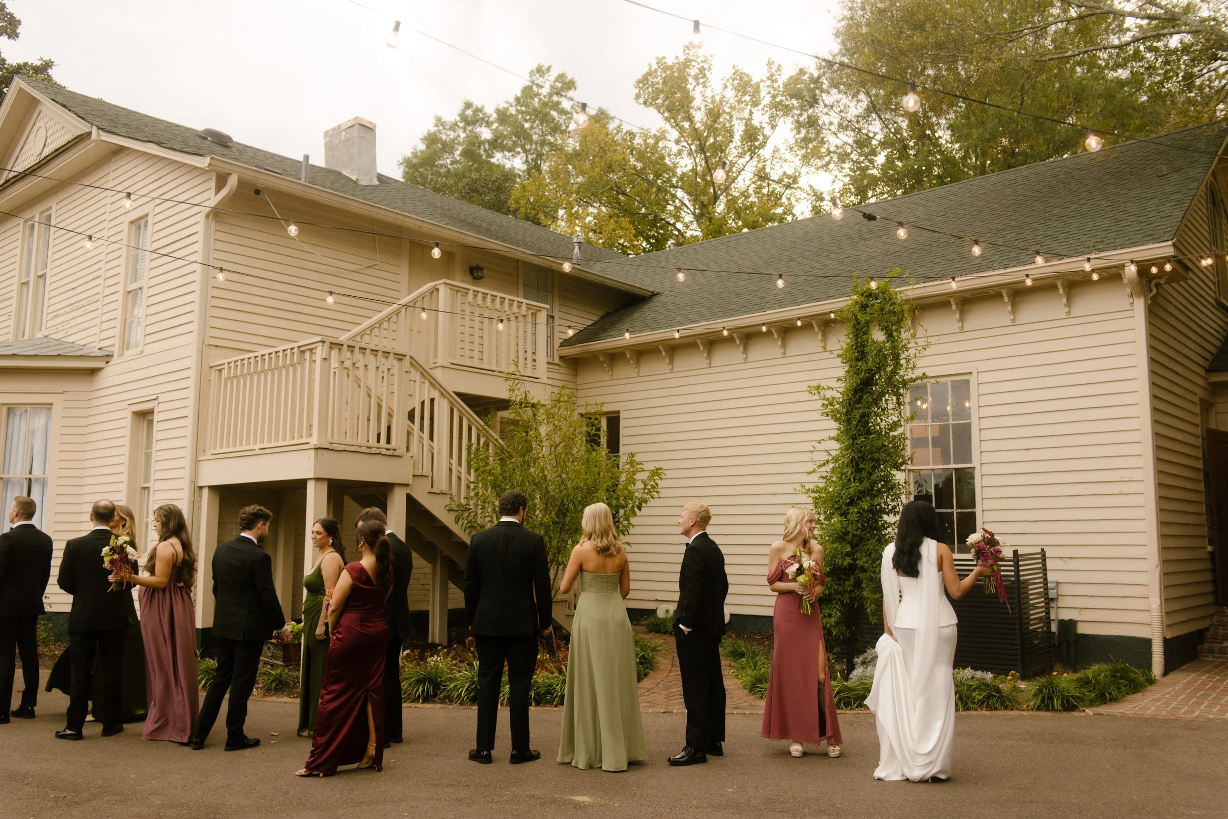 Outdoor wedding cocktail hour with guests mingling beneath string lights and bridesmaids in moody wedding colors.