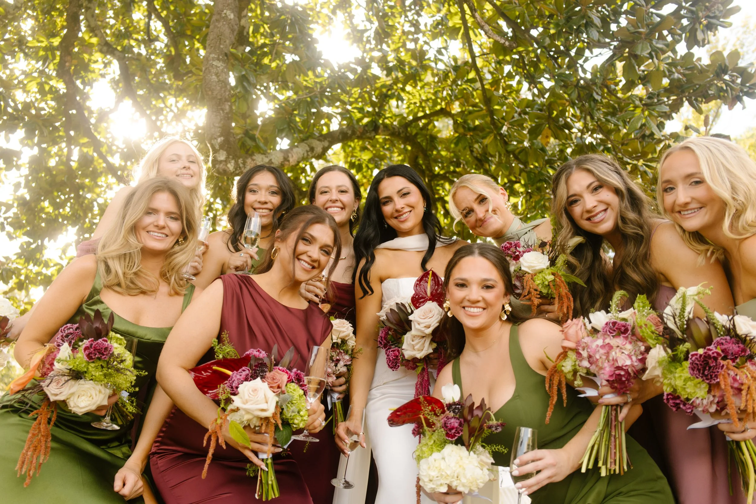 Bride and bridesmaids smiling together with lush bouquets featuring moody wedding colors of burgundy, plum, olive green, and ivory.