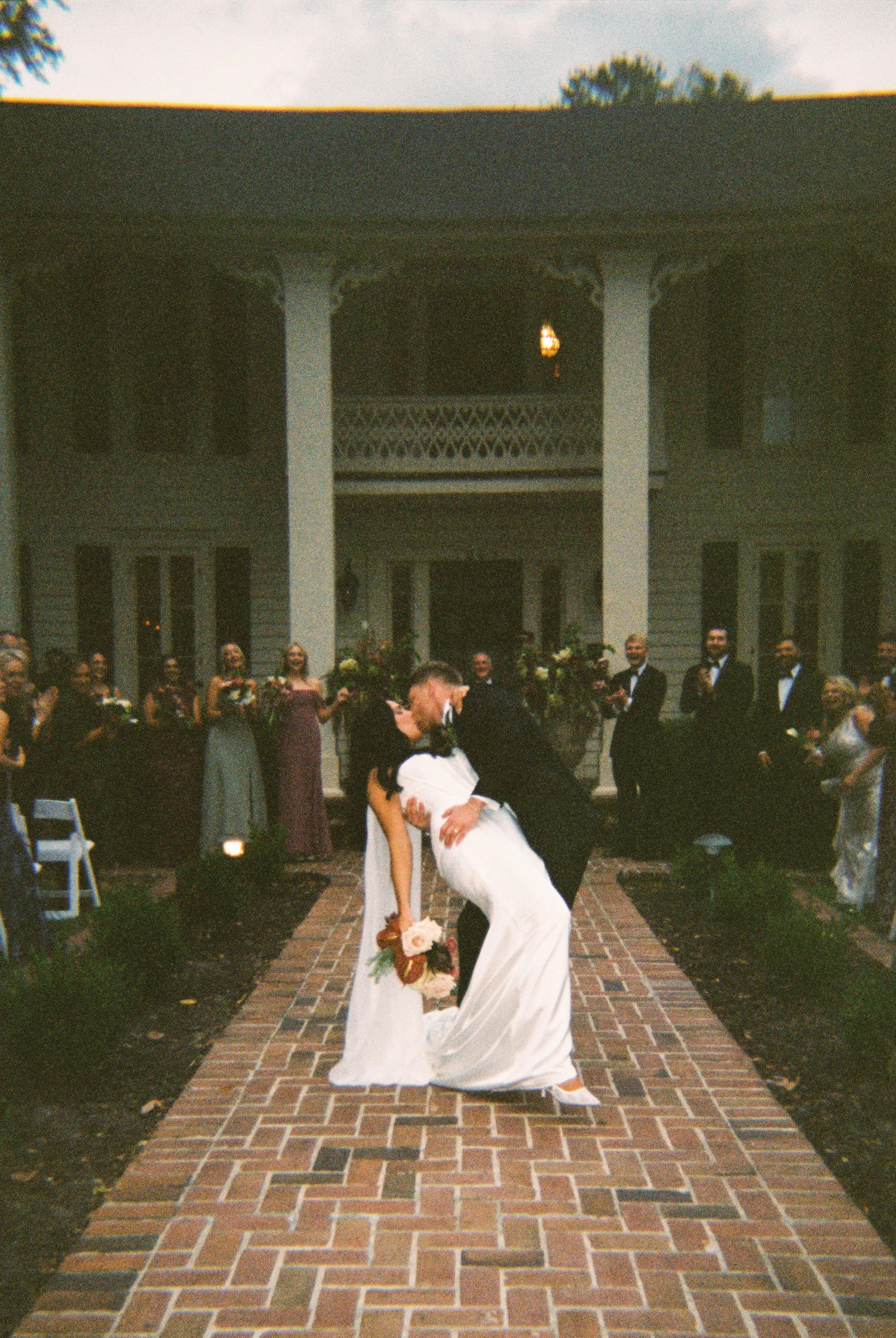 Film wedding photograph of a bride and groom sharing a dip kiss in front of their wedding party at an outdoor ceremony.