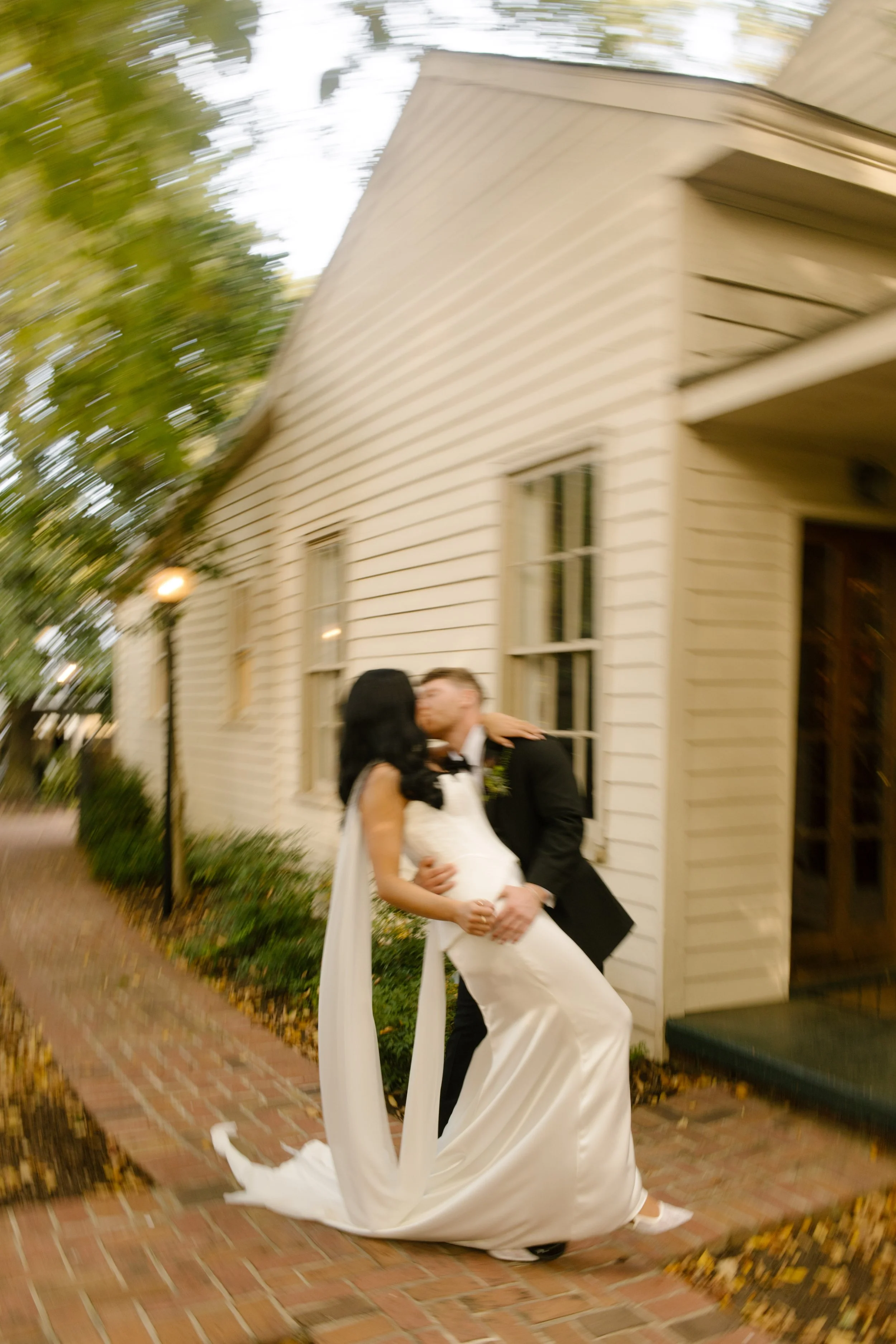 Bride and groom sharing a romantic dip kiss on a brick walkway outside a historic white wedding venue.