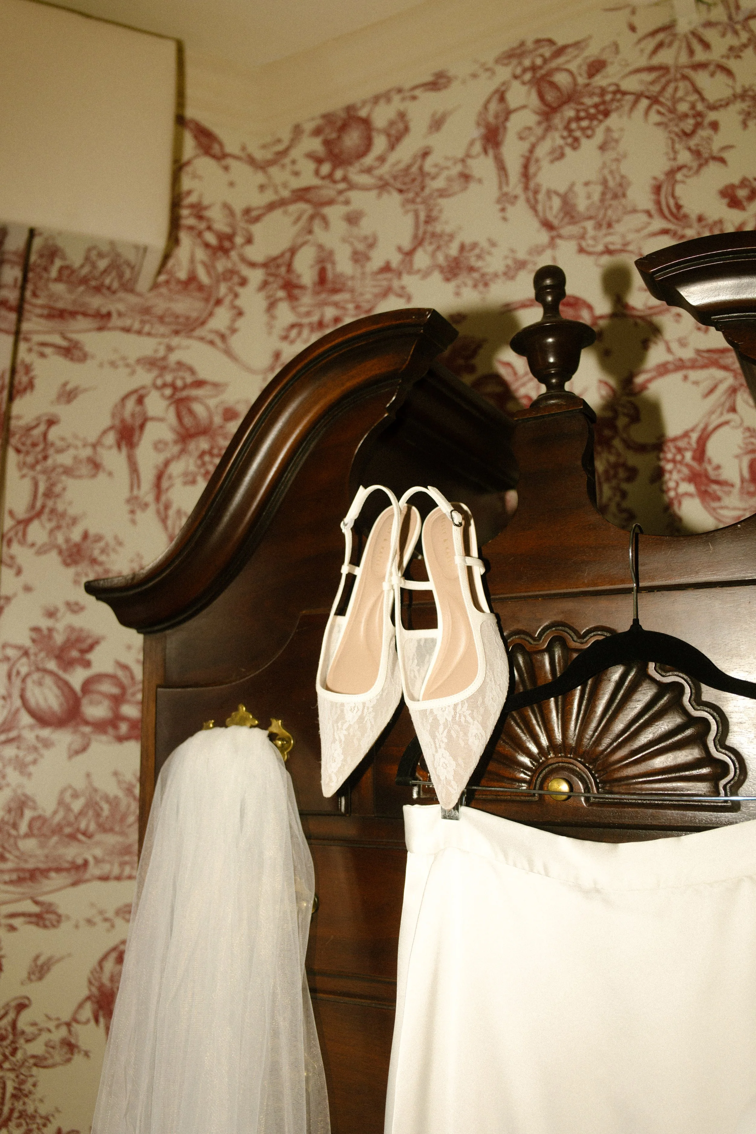 Bridal details featuring moody wedding colors with white lace heels, a veil, and a dark wooden bed frame against red toile wallpaper.