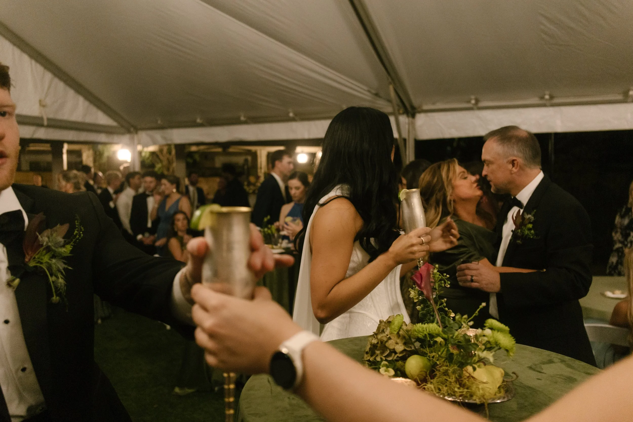 Candid wedding reception with guests gathered around a green table featuring moody wedding colors and lush floral arrangements.