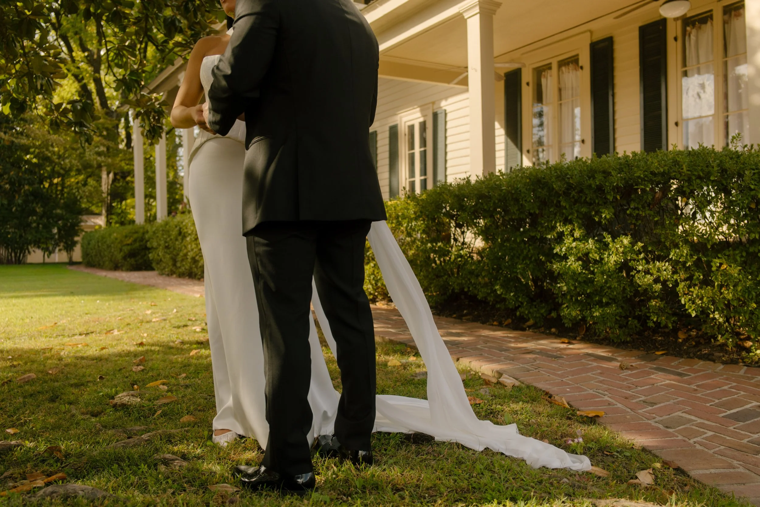 Close-up of a bride and groom embracing outdoors, with her long white veil trailing across the grass beside a historic venue.