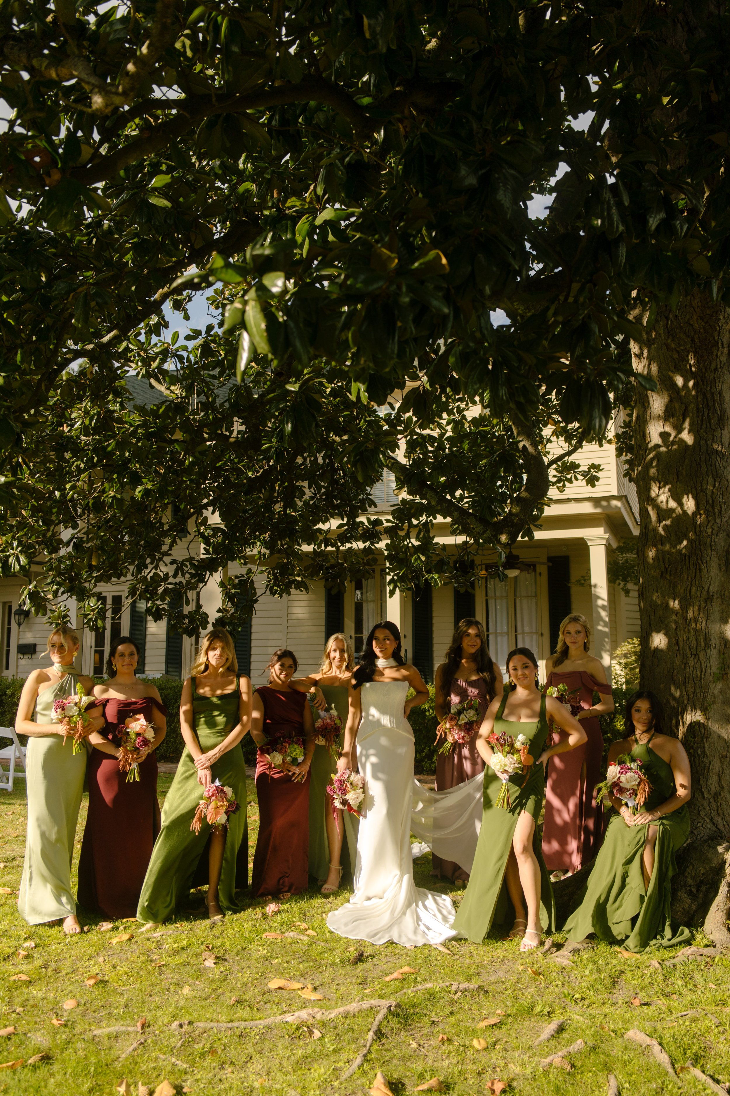 Bride and bridesmaids posing beneath a large tree in dresses featuring moody wedding colors of burgundy, olive green, and mauve.