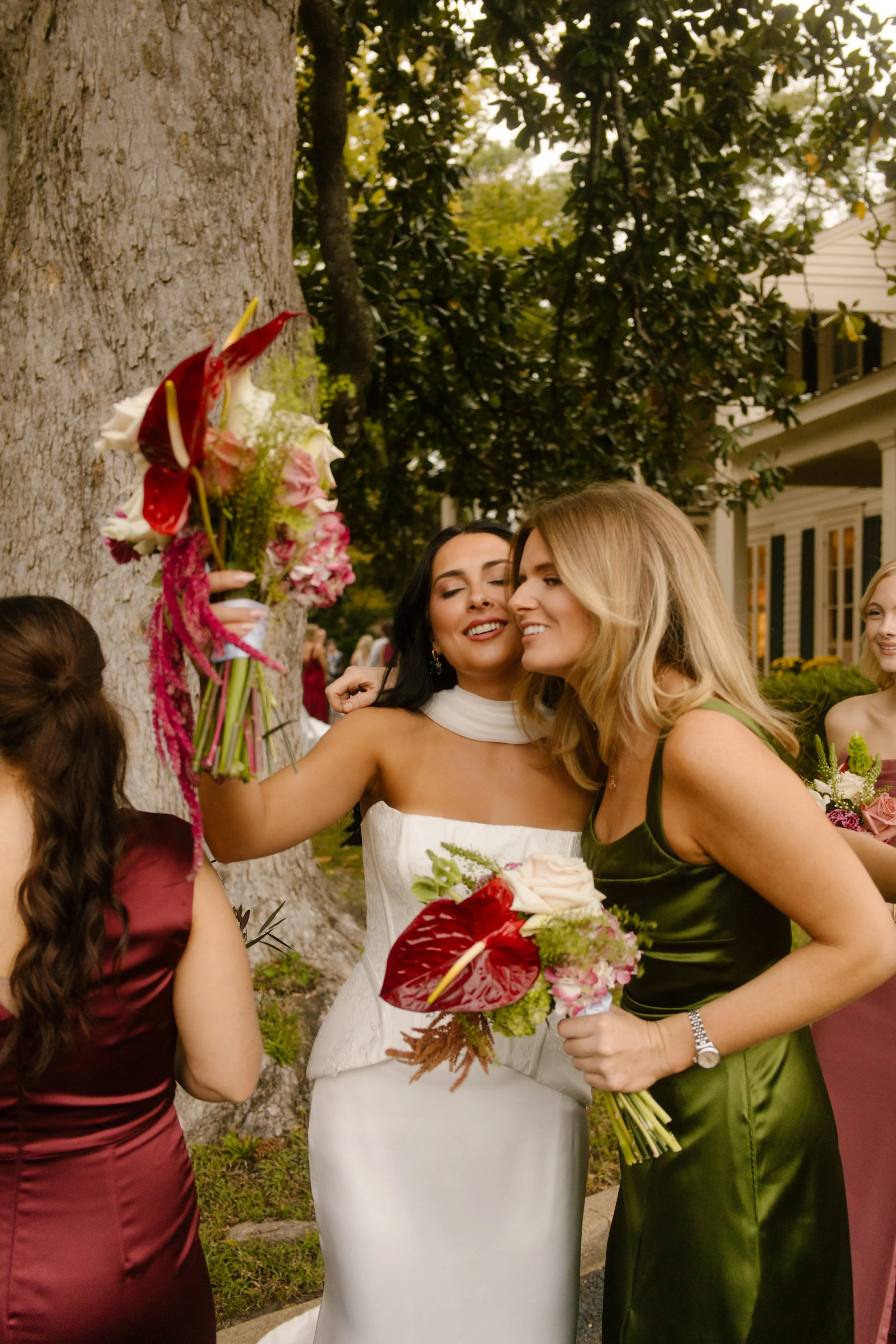 Bride smiling with a bridesmaid in an olive green satin dress while holding dramatic red and pink wedding florals.