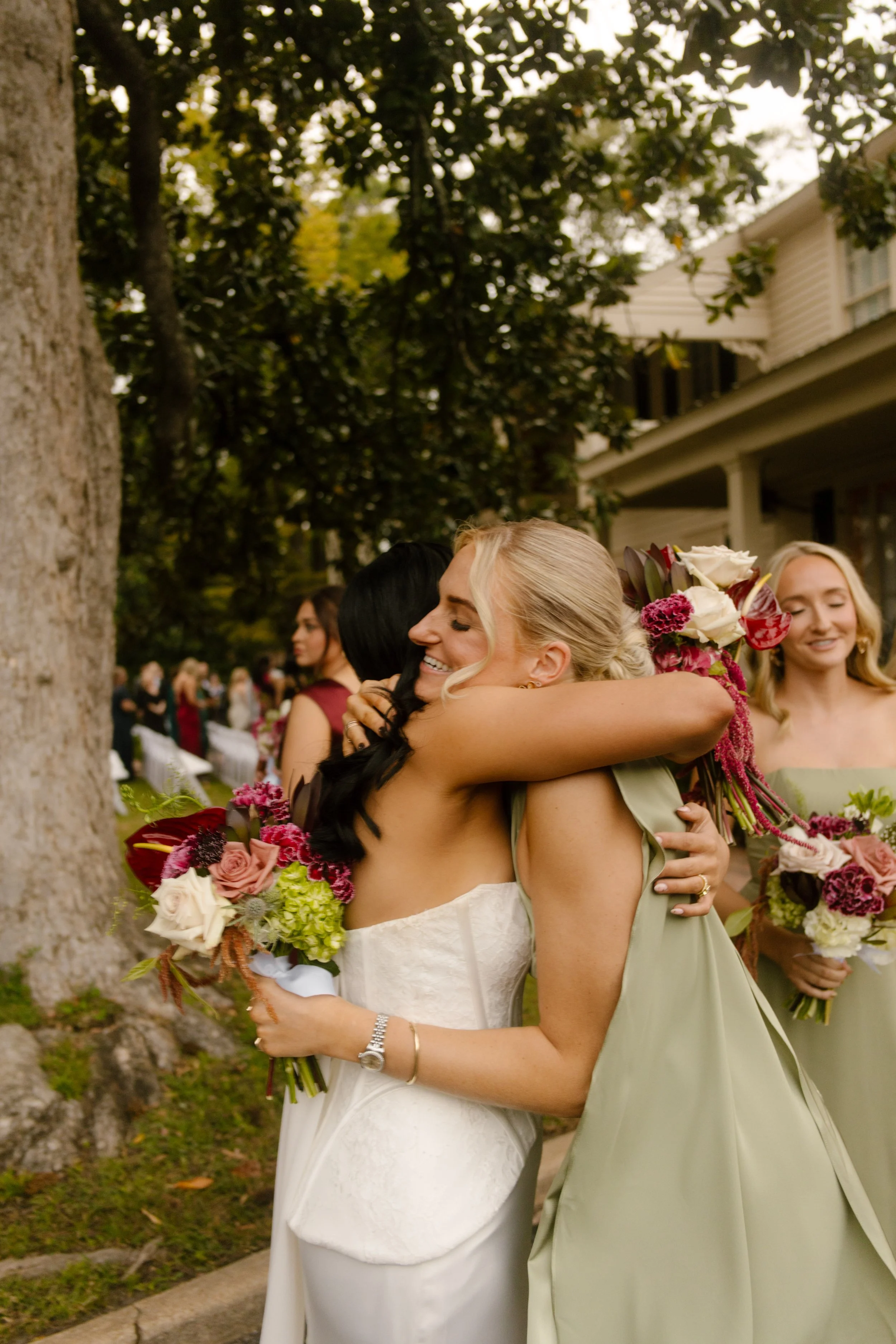 Bride hugging a bridesmaid in a sage green dress while holding a bouquet featuring moody wedding colors of burgundy, pink, and ivory.