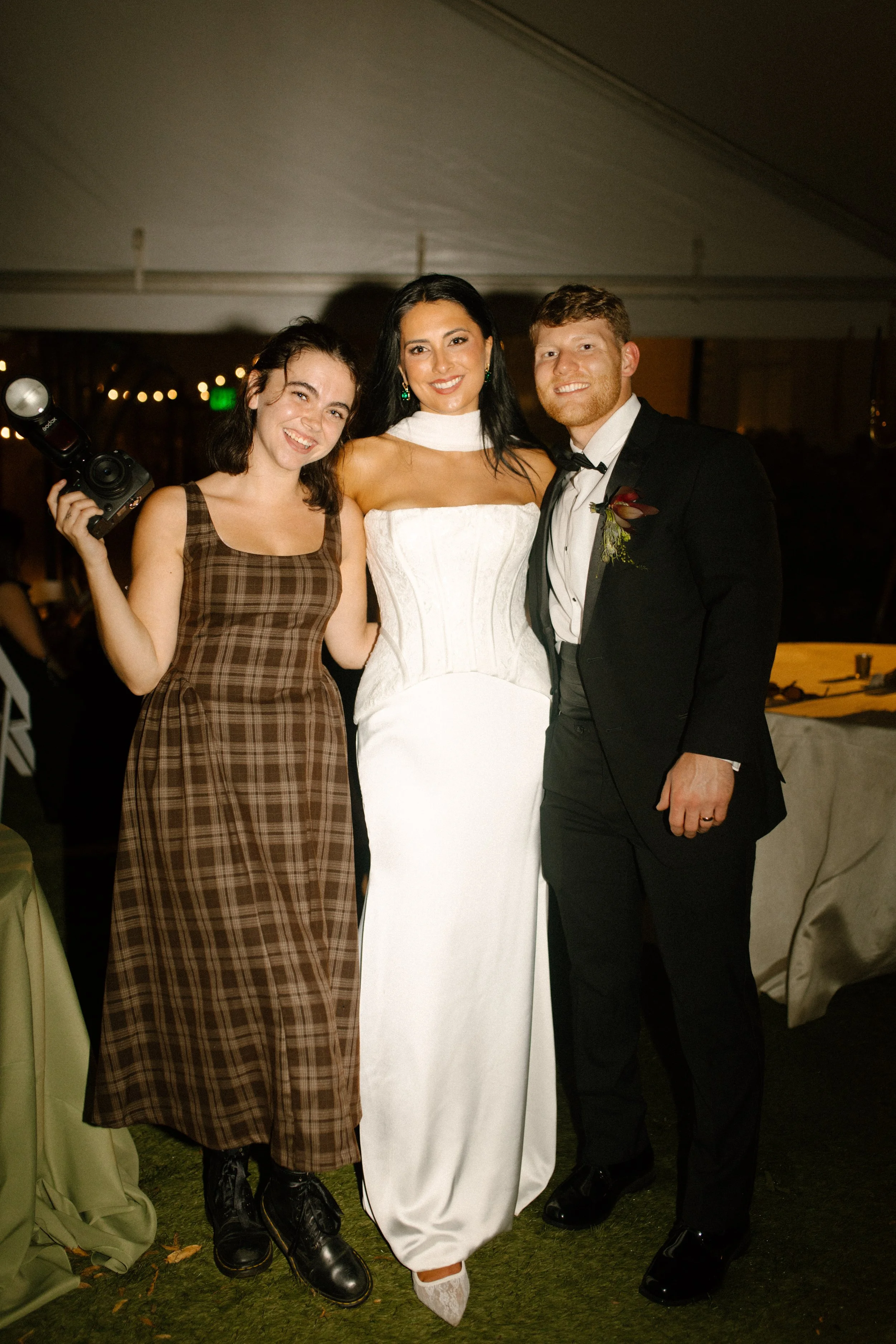 Bride and groom posing with their wedding photographer at a nighttime reception beneath a tent.