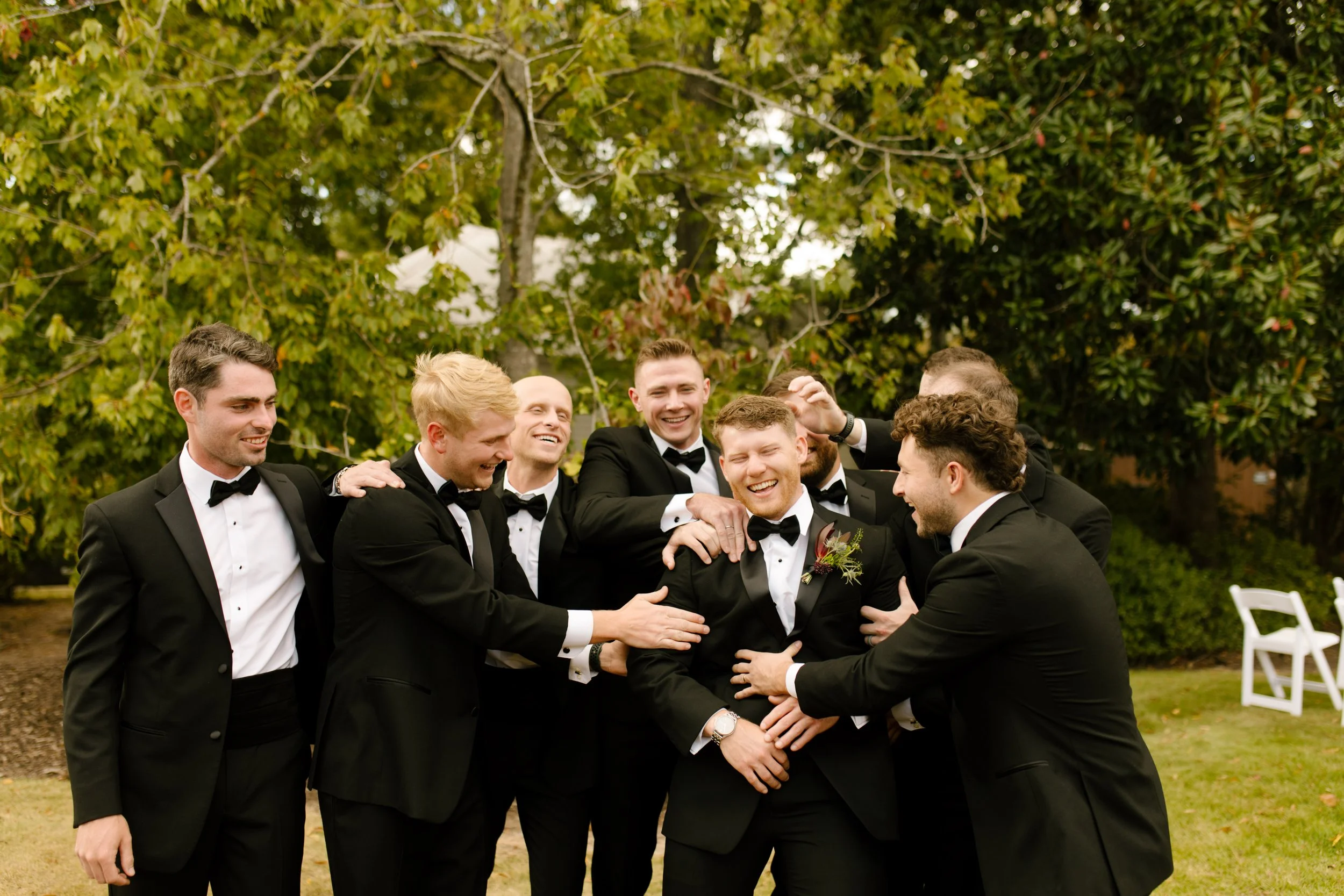 Groom laughing with his groomsmen in black tuxedos during a candid outdoor wedding party photo.