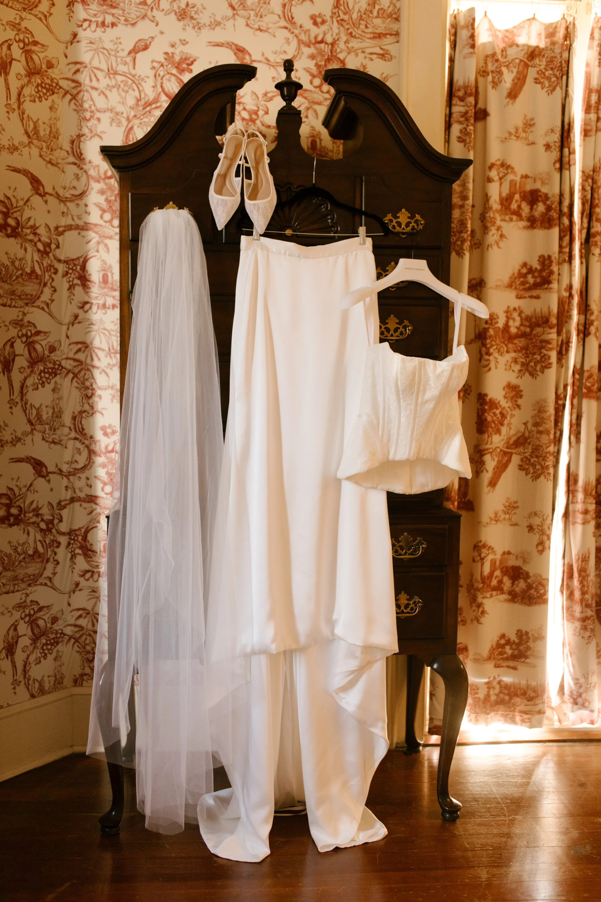 Bridal details featuring a white corset wedding gown, pointed-toe heels, and a long veil displayed against vintage red toile wallpaper.