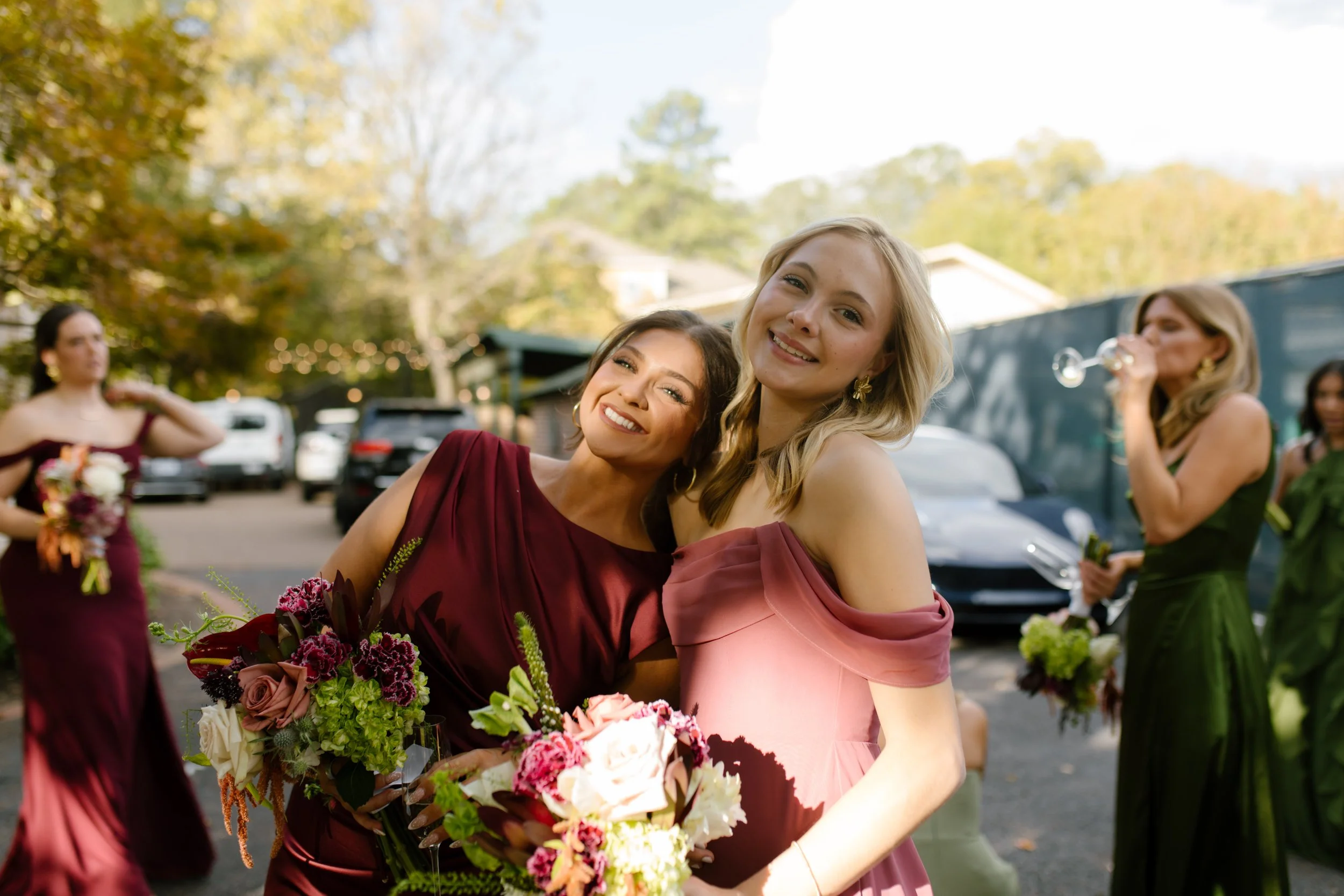 Two bridesmaids smiling together in burgundy and dusty rose dresses while holding colorful wedding bouquets.