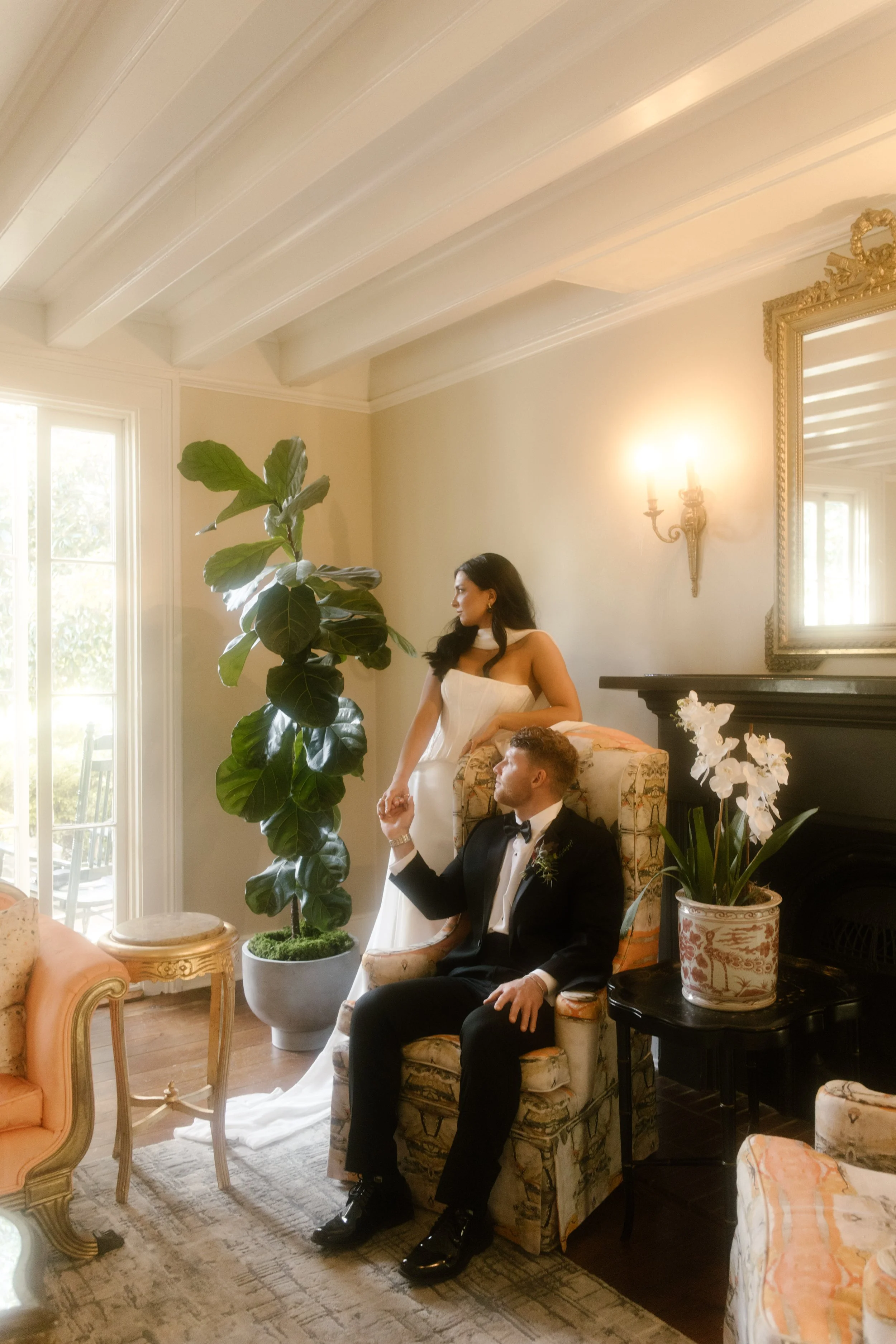 Bride and groom posing in a sunlit vintage sitting room with warm lighting, antique furnishings, and moody wedding colors.