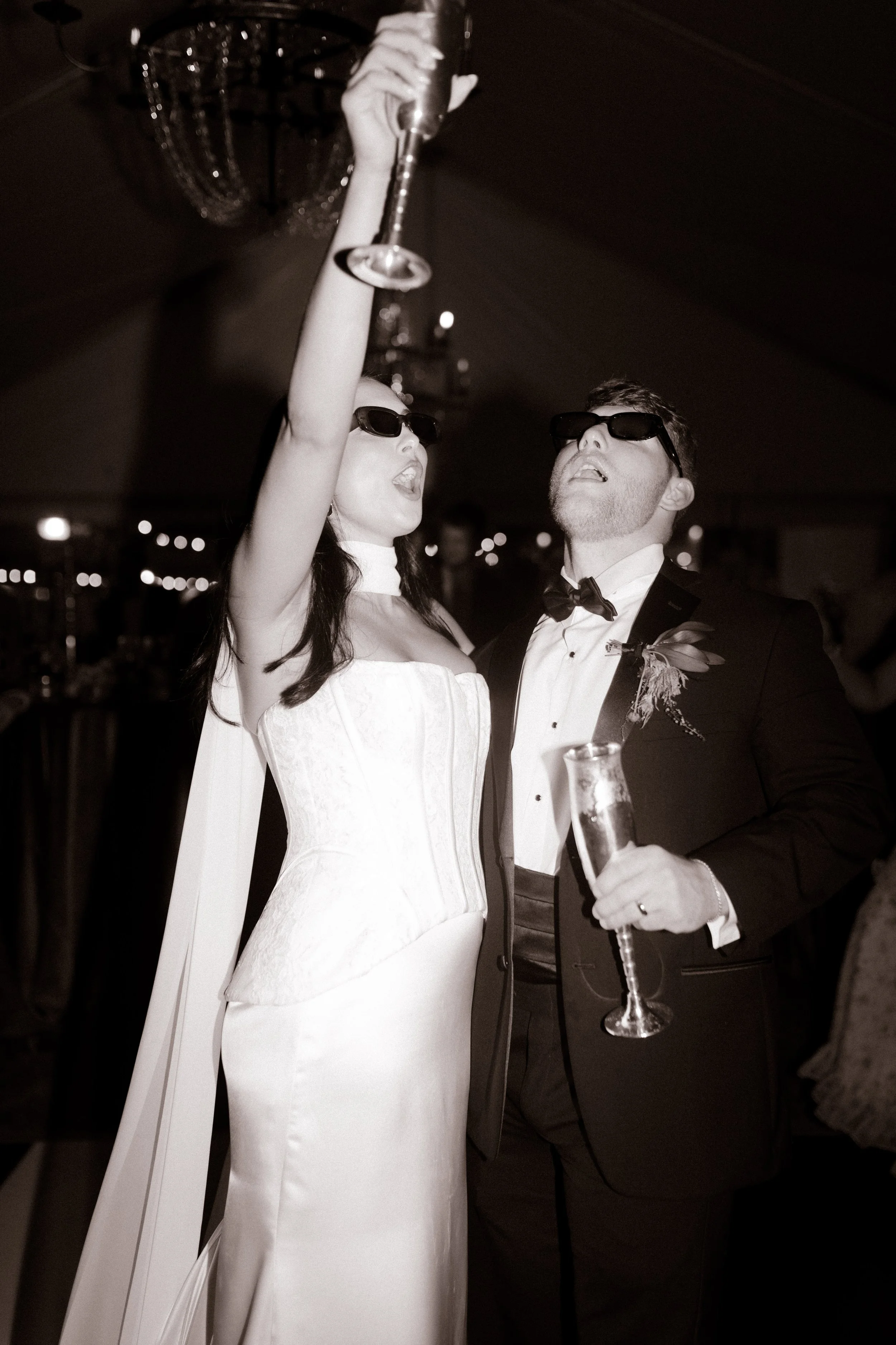 Black-and-white wedding reception photo of the bride and groom wearing sunglasses and raising champagne glasses on the dance floor.