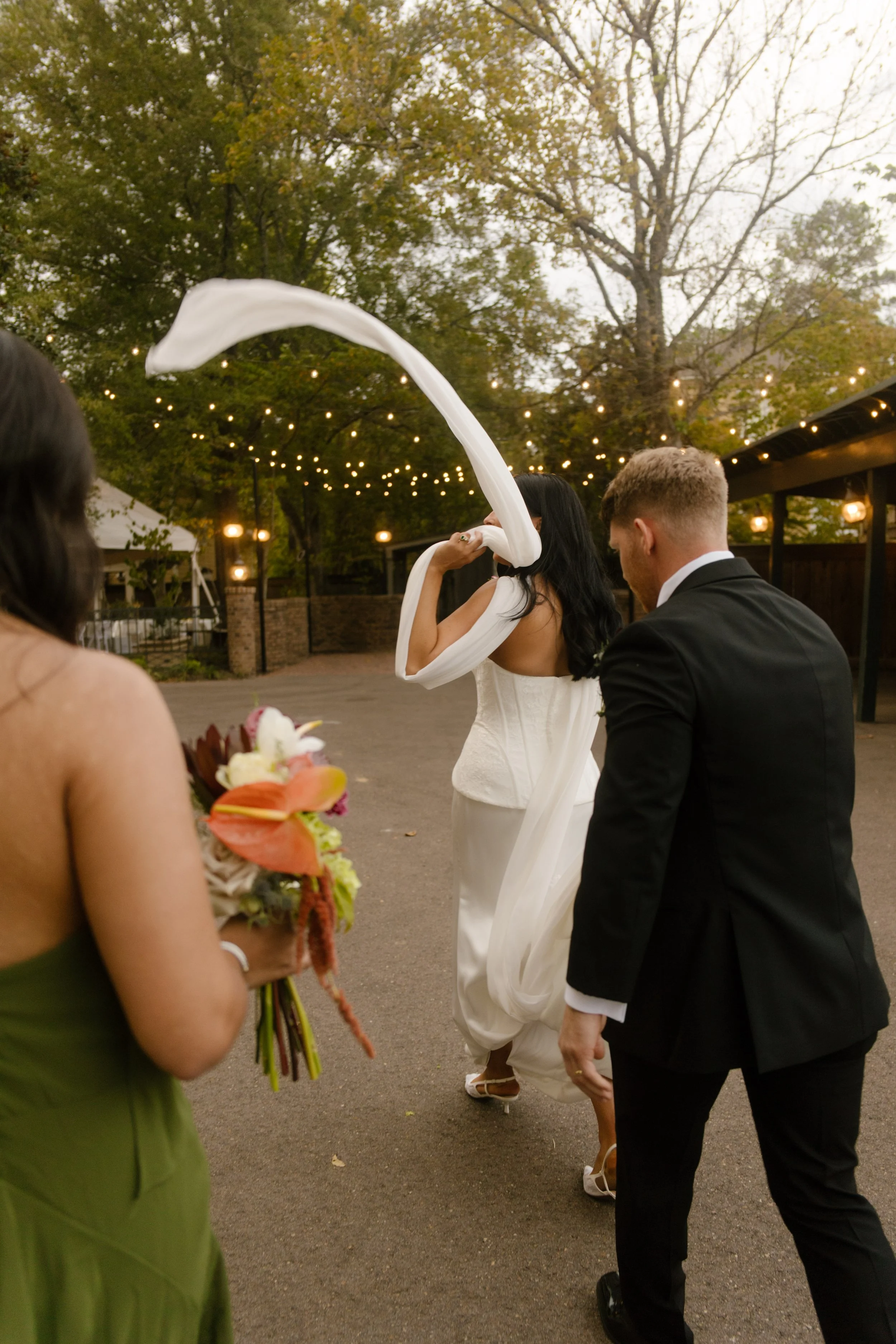 Bride and groom walking toward their wedding reception beneath string lights, with a bridesmaid carrying a colorful bouquet featuring moody wedding colors.