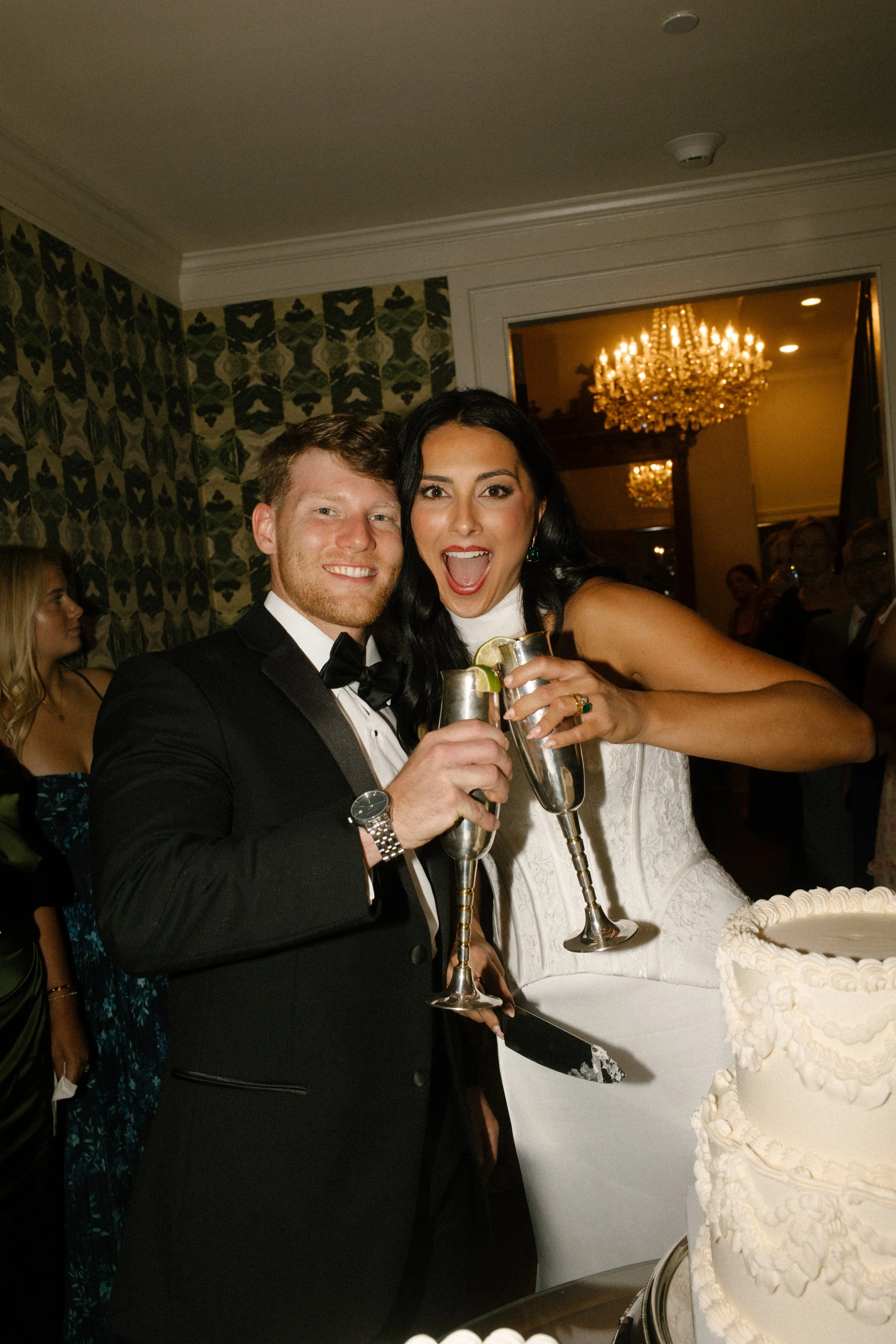 Bride and groom celebrating beside their wedding cake, with moody wedding colors in the green patterned wallpaper and warm reception lighting.