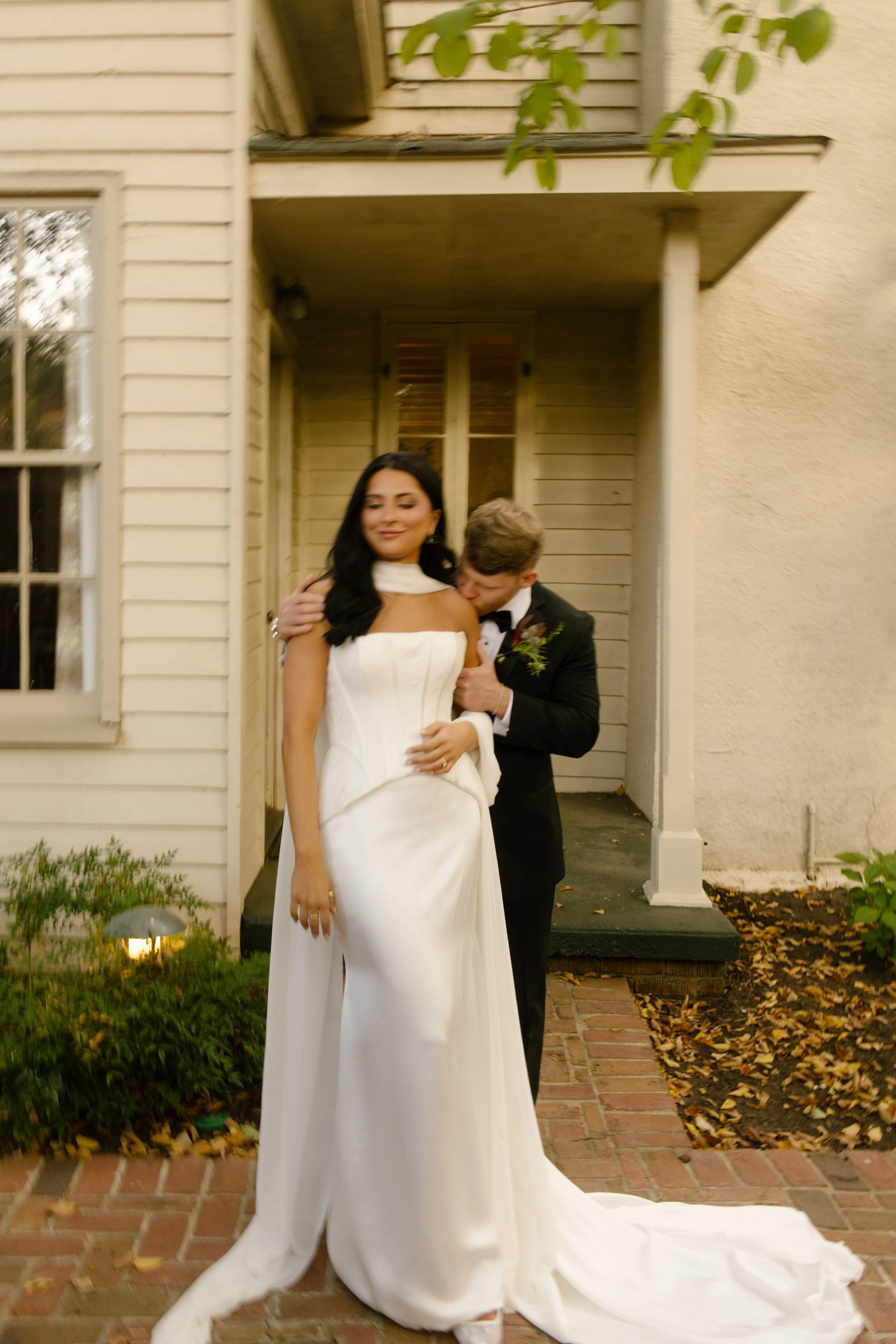 Bride and groom sharing a romantic outdoor portrait beside a historic wedding venue, with her long white veil trailing across the brick walkway.