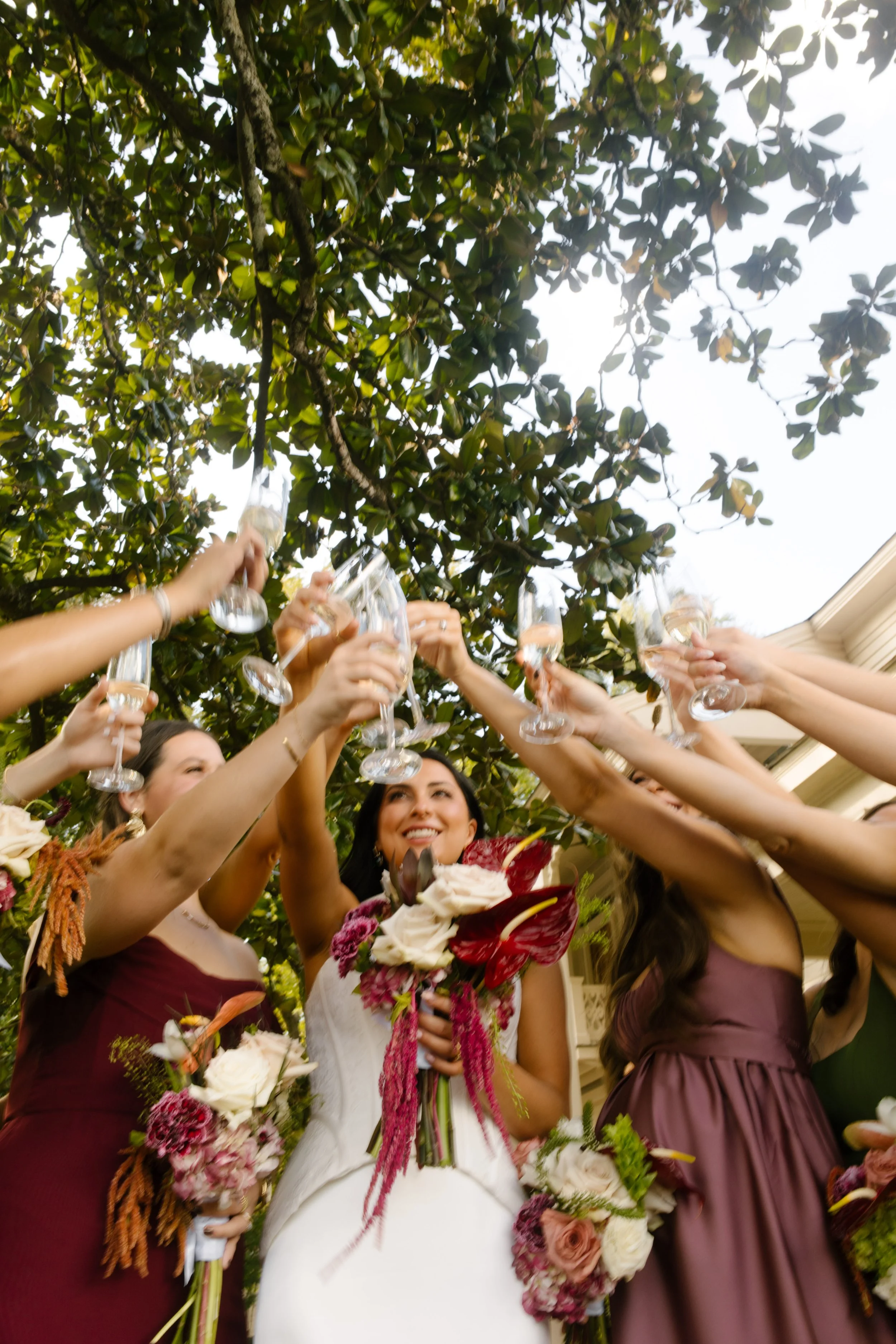 Bride and bridesmaids raising champagne glasses beneath the trees, wearing moody wedding colors of burgundy, mauve, and olive green.