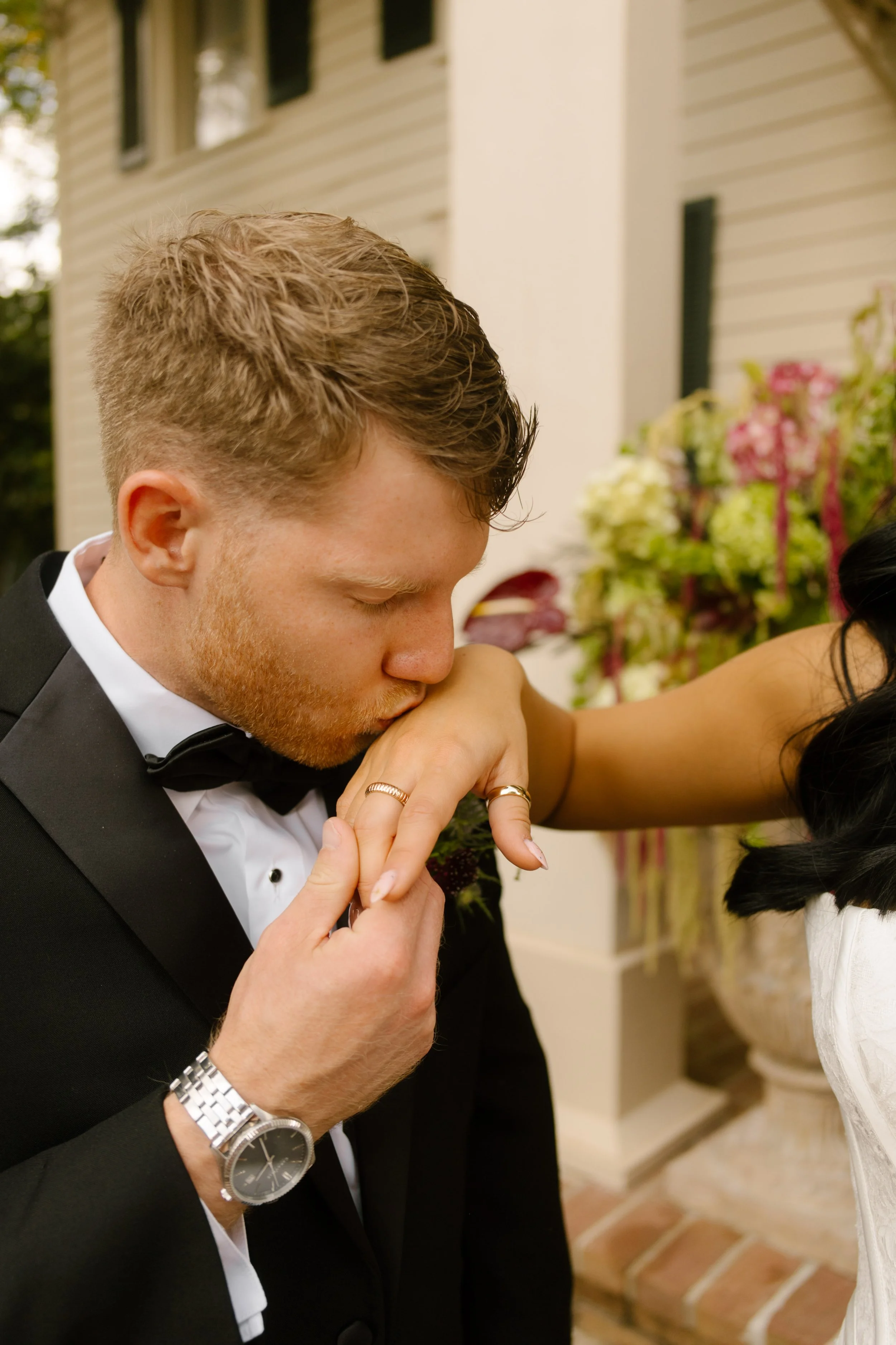 Groom kissing the bride's hand during a romantic wedding moment with burgundy and green floral arrangements in the background.