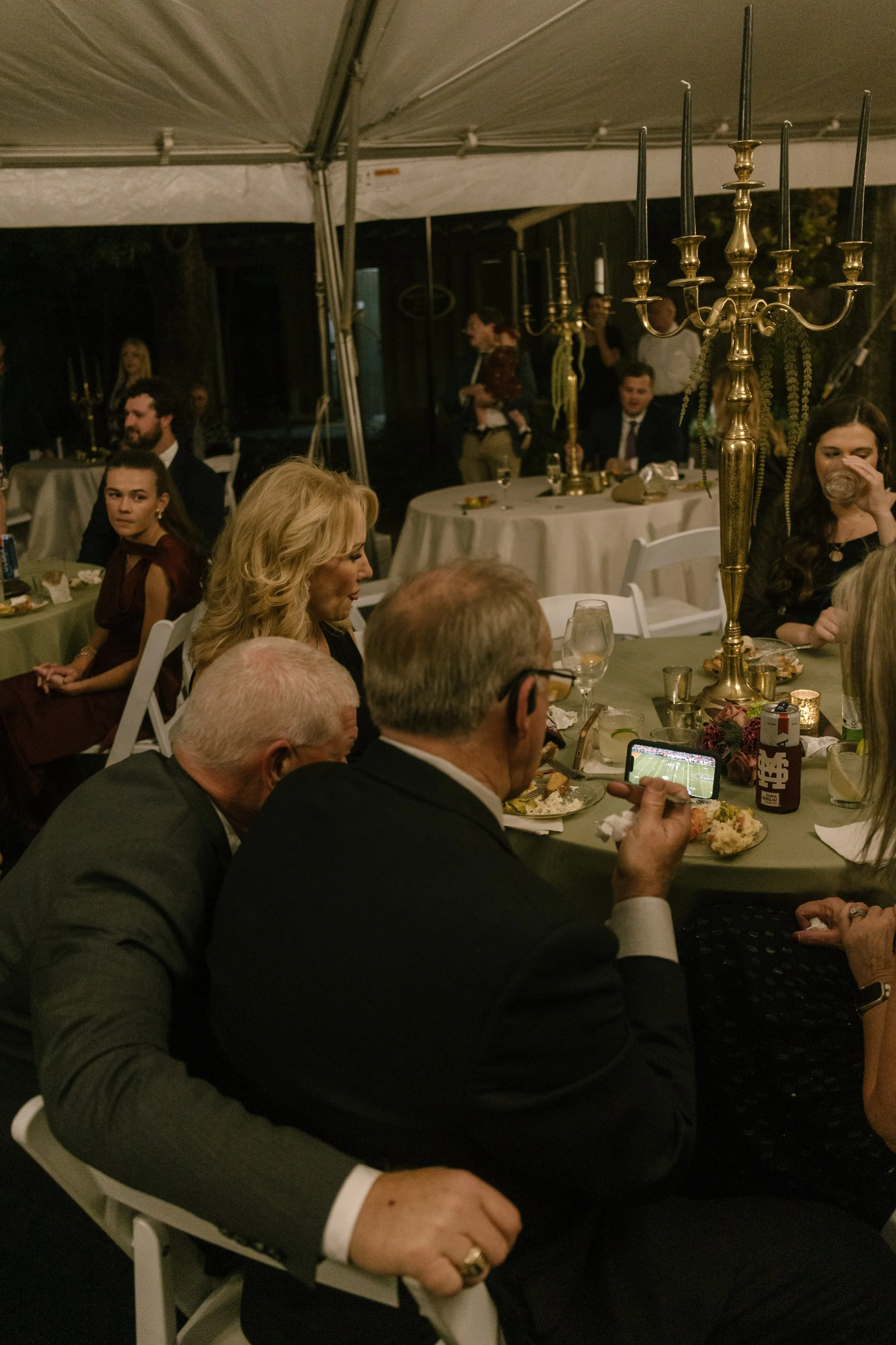 Wedding guests seated beneath a reception tent with antique brass candelabras, green table linens, and moody wedding colors.