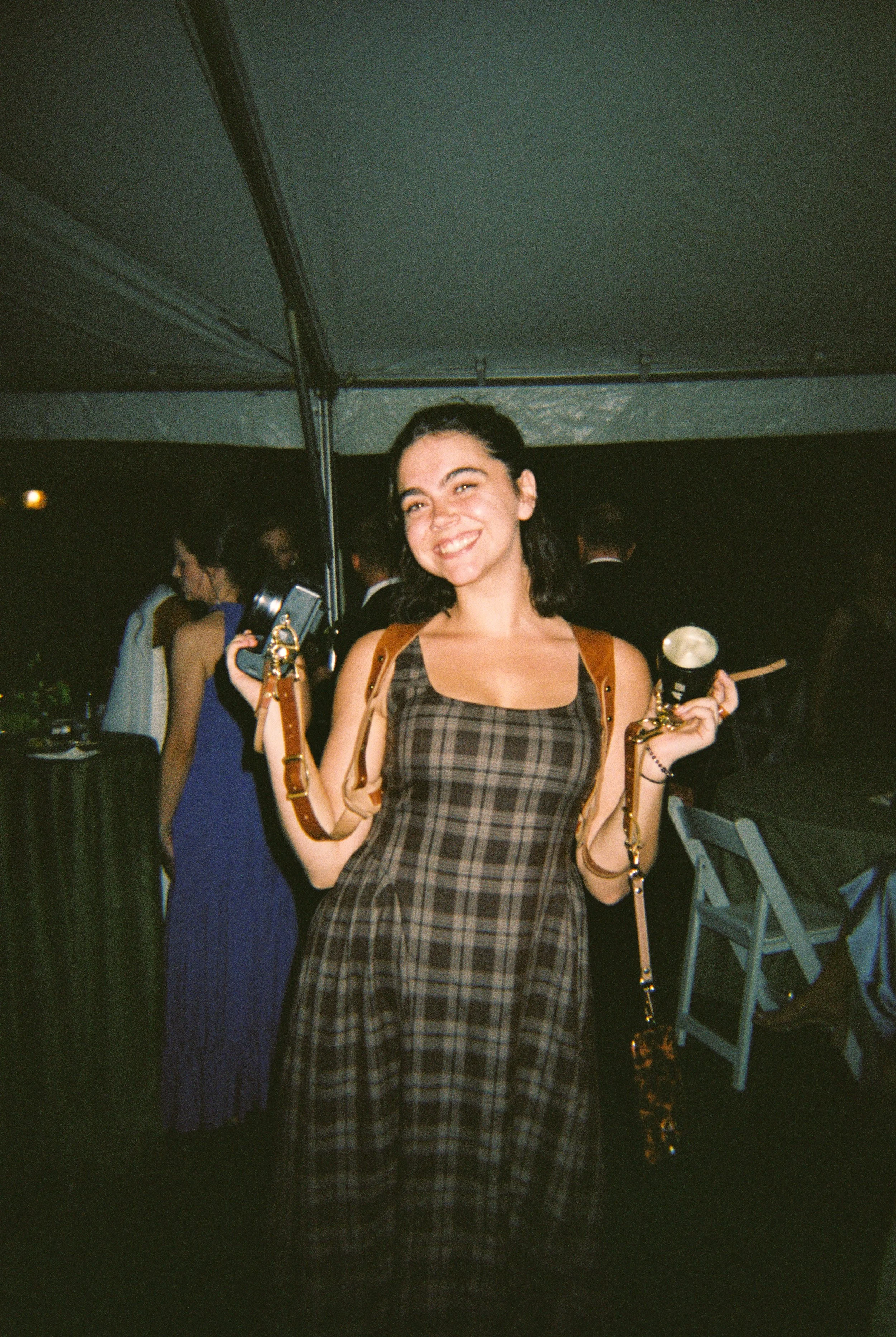 Wedding photographer smiling with her cameras during an evening reception, wearing a plaid dress in moody wedding colors.